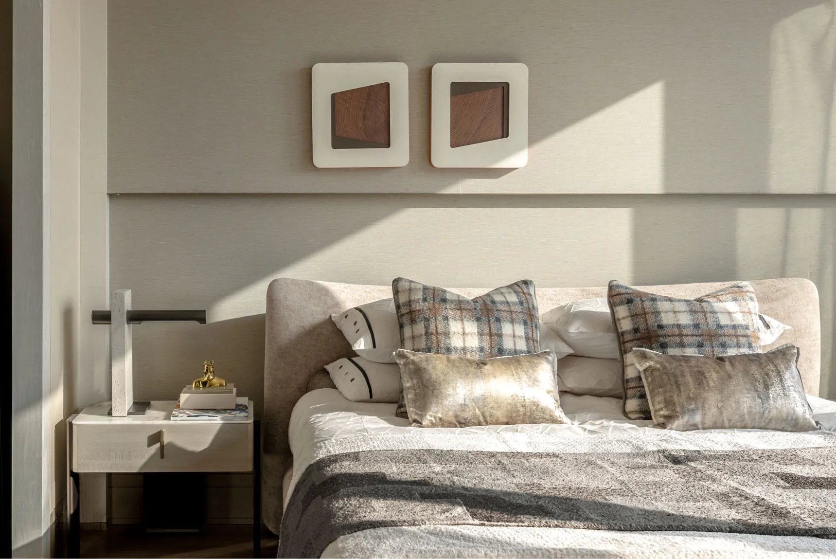 A modern bedroom featuring neutral textiles on the bed and a side table with a lamp and books.