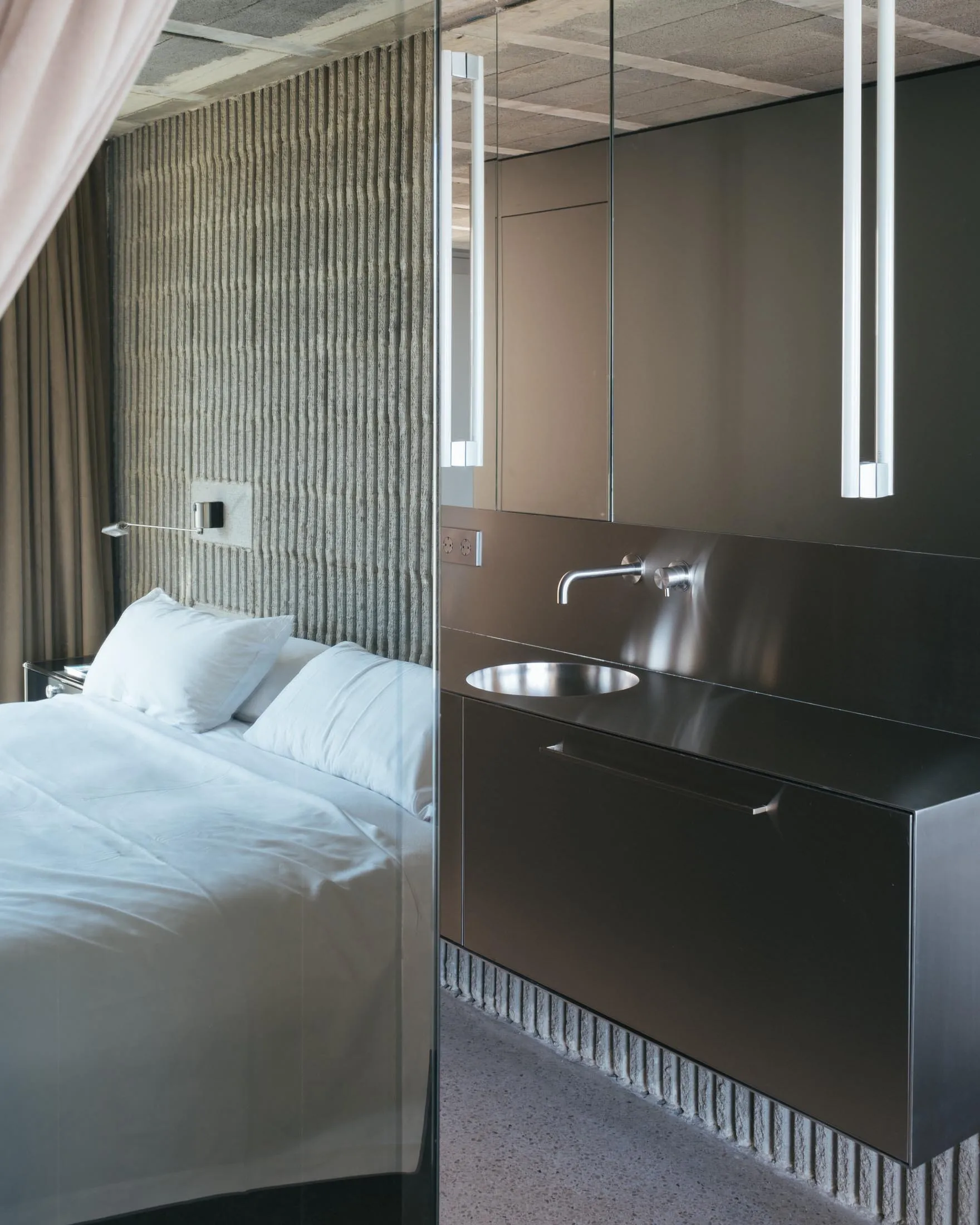 Modern minimalist bedroom featuring a metal vanity sink, white linens, and a textured wall behind the bed.