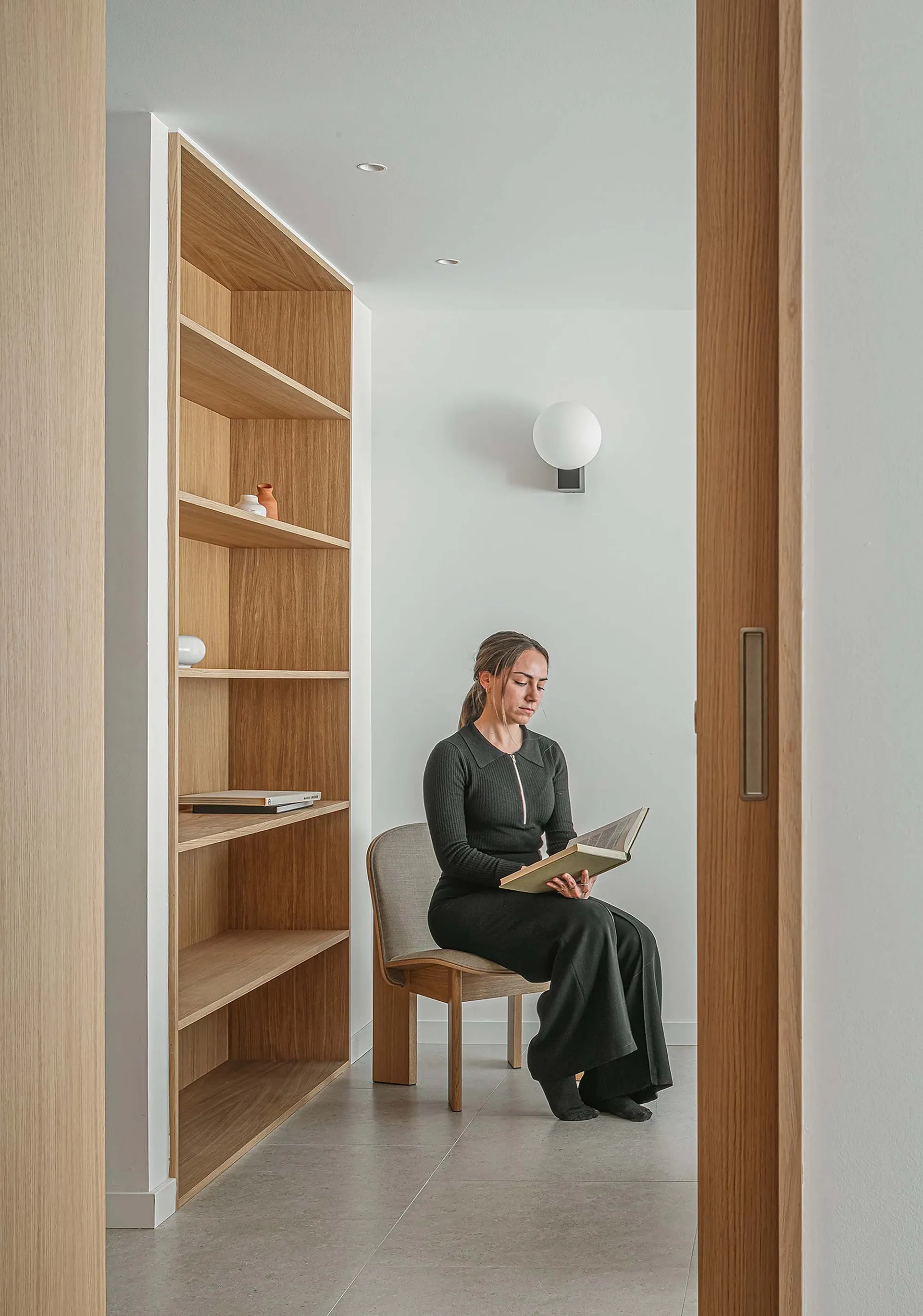 A woman sitting on a wooden chair in a minimalist library, holding a book.