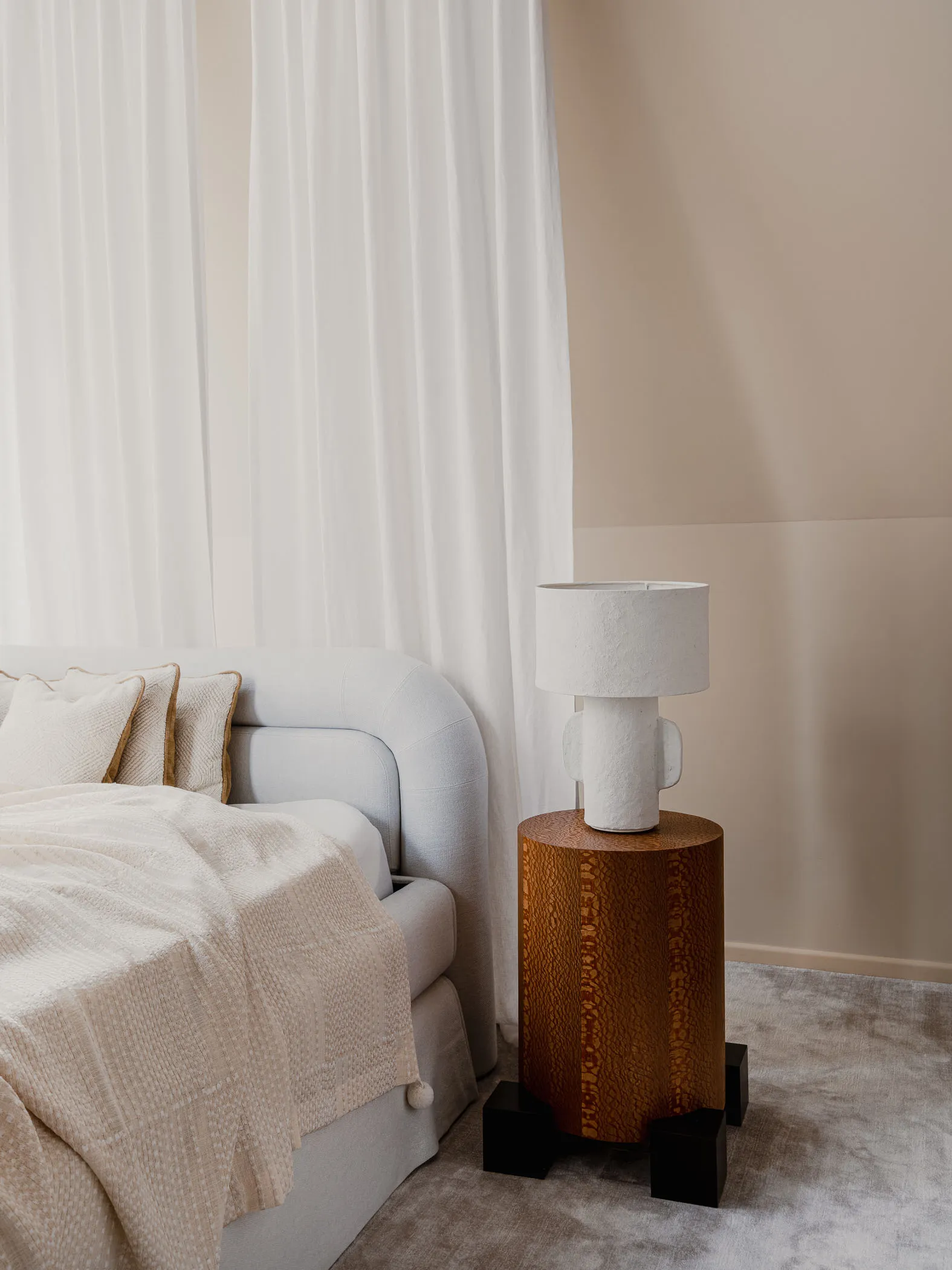 A minimalist bedroom featuring a white bed with pillows, a beige blanket, and a wooden side table with a ceramic lamp. White