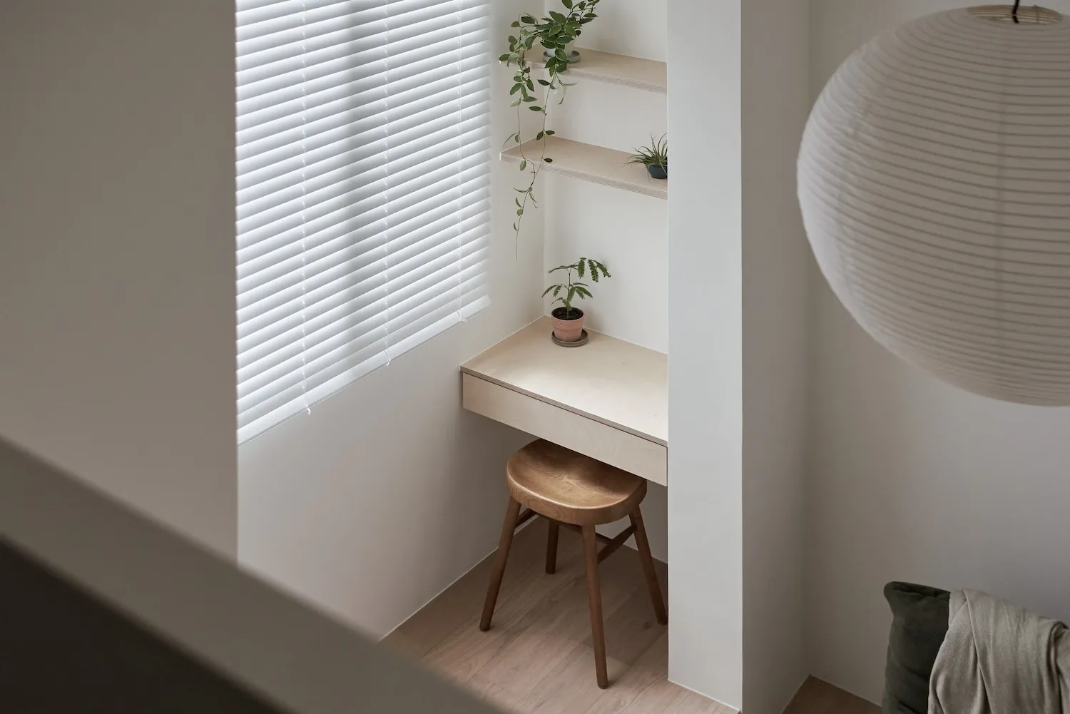 A minimalist interior design featuring a wooden desk, stool, and plants in front of a window with white blinds.