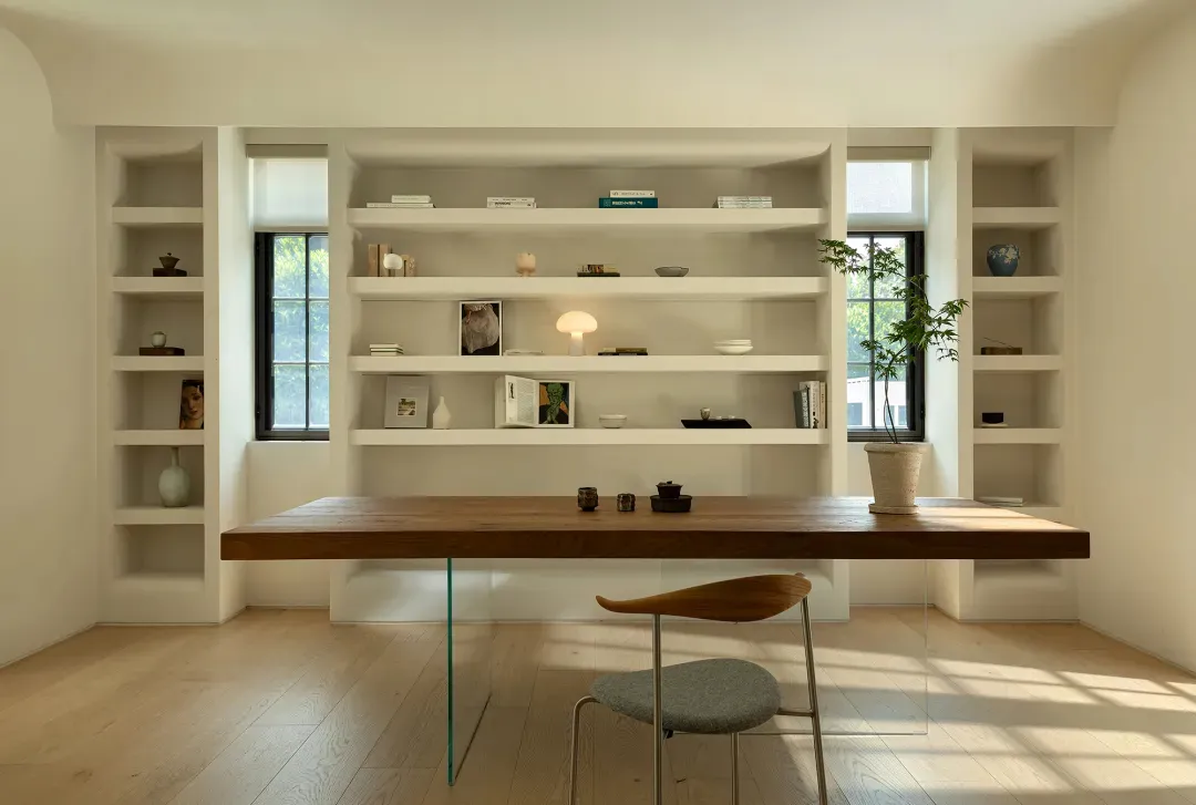 A modern home office with a wooden desk, glass table legs, and built-in shelves.