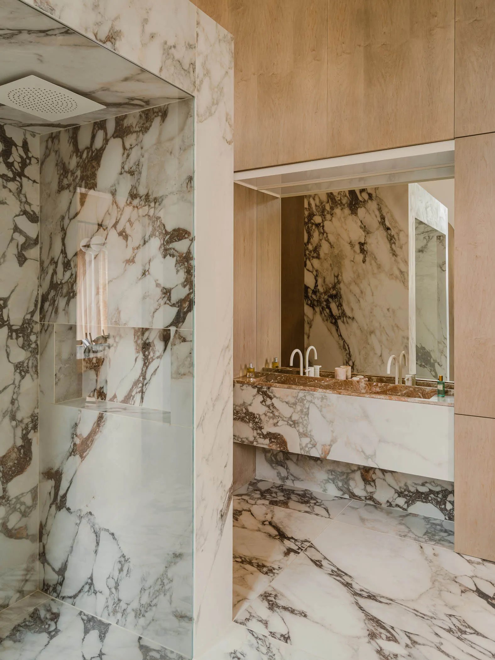 A modern bathroom featuring marble walls and flooring, with a double sink vanity and glass shower enclosure.