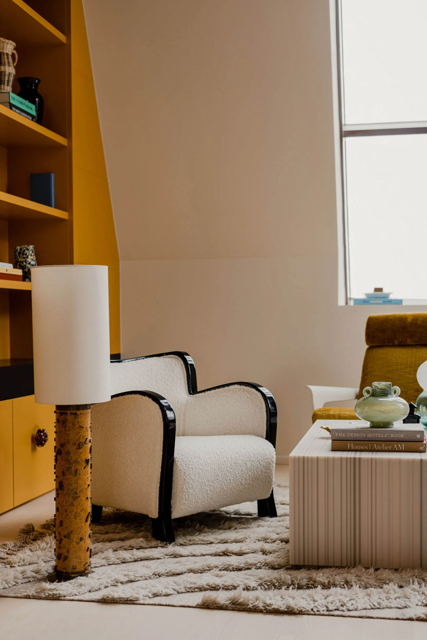 A modern residential living room featuring a textured fabric armchair, a coffee table with books and a vase, and a yellow boo