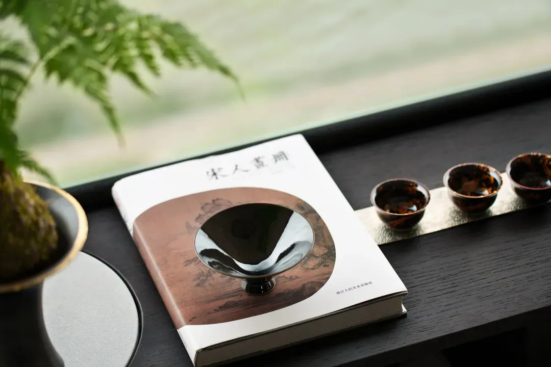 A photograph of a ceramic sake cup, a book titled 'Sōren' featuring artwork, and black and gold ceramic cups arranged on a wo
