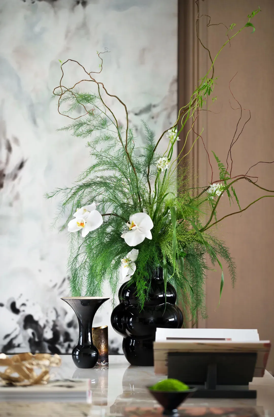 A black vase with white orchids, greenery, and spiral branches on a reflective surface.