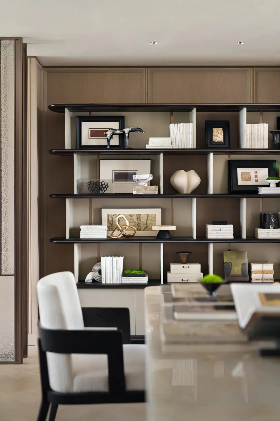 A modern, minimalist library interior featuring a shelf with books, frames, and decorative objects.