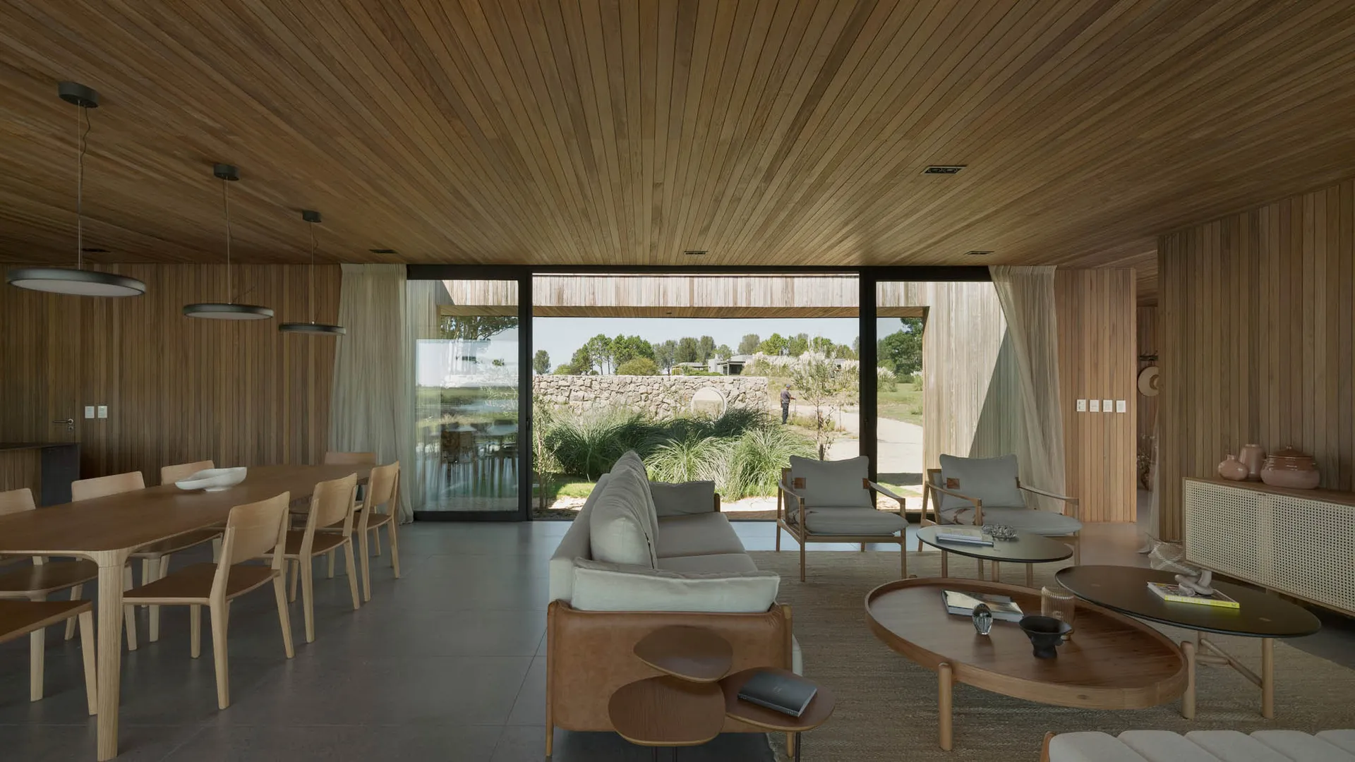 Modern minimalist living room with wooden coffee tables, leather sofa, and sliding glass doors leading to an outdoor area.