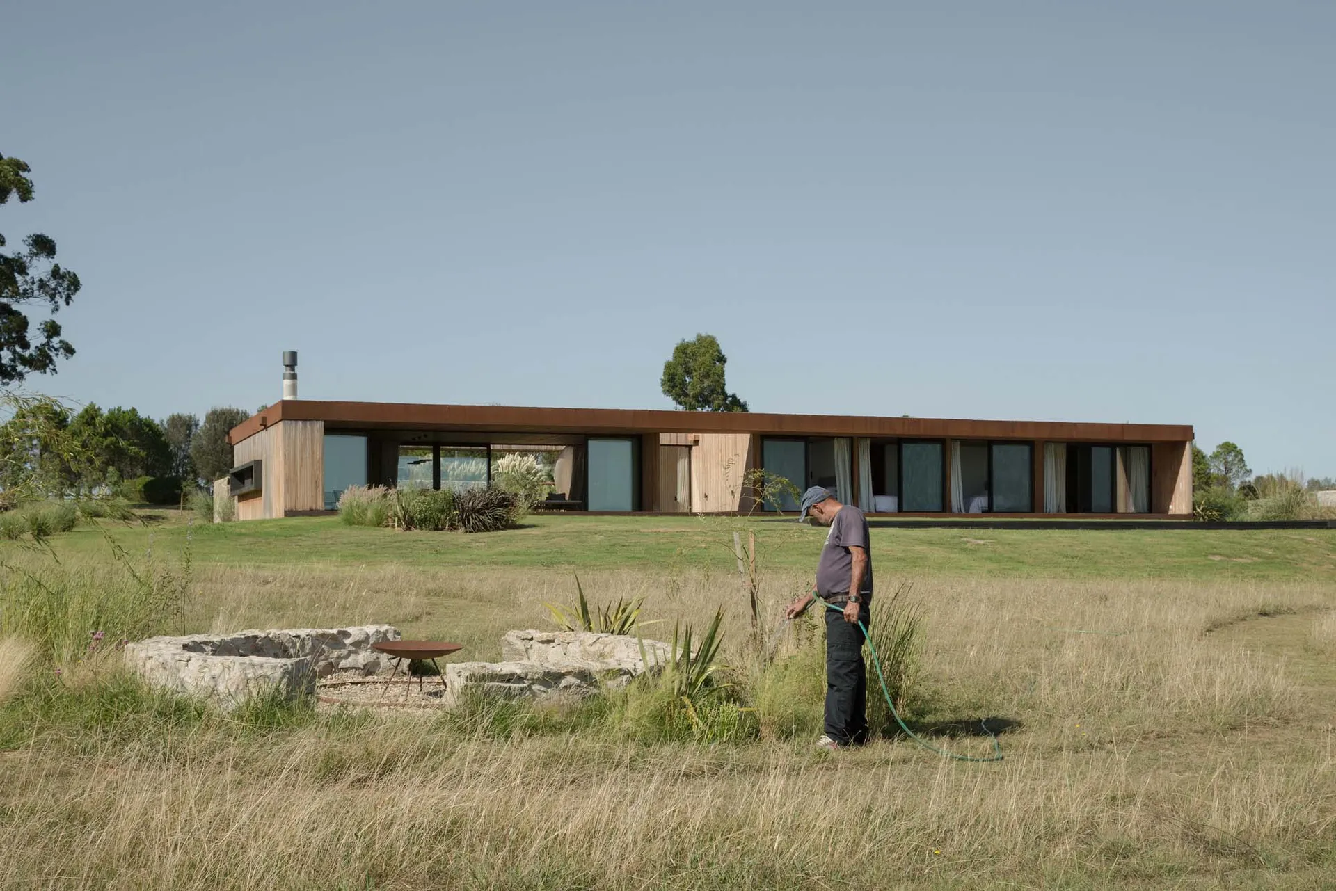 Man watering lawn on a sunny day in front of a modern residential exterior with a metal roof and metal roof vent.