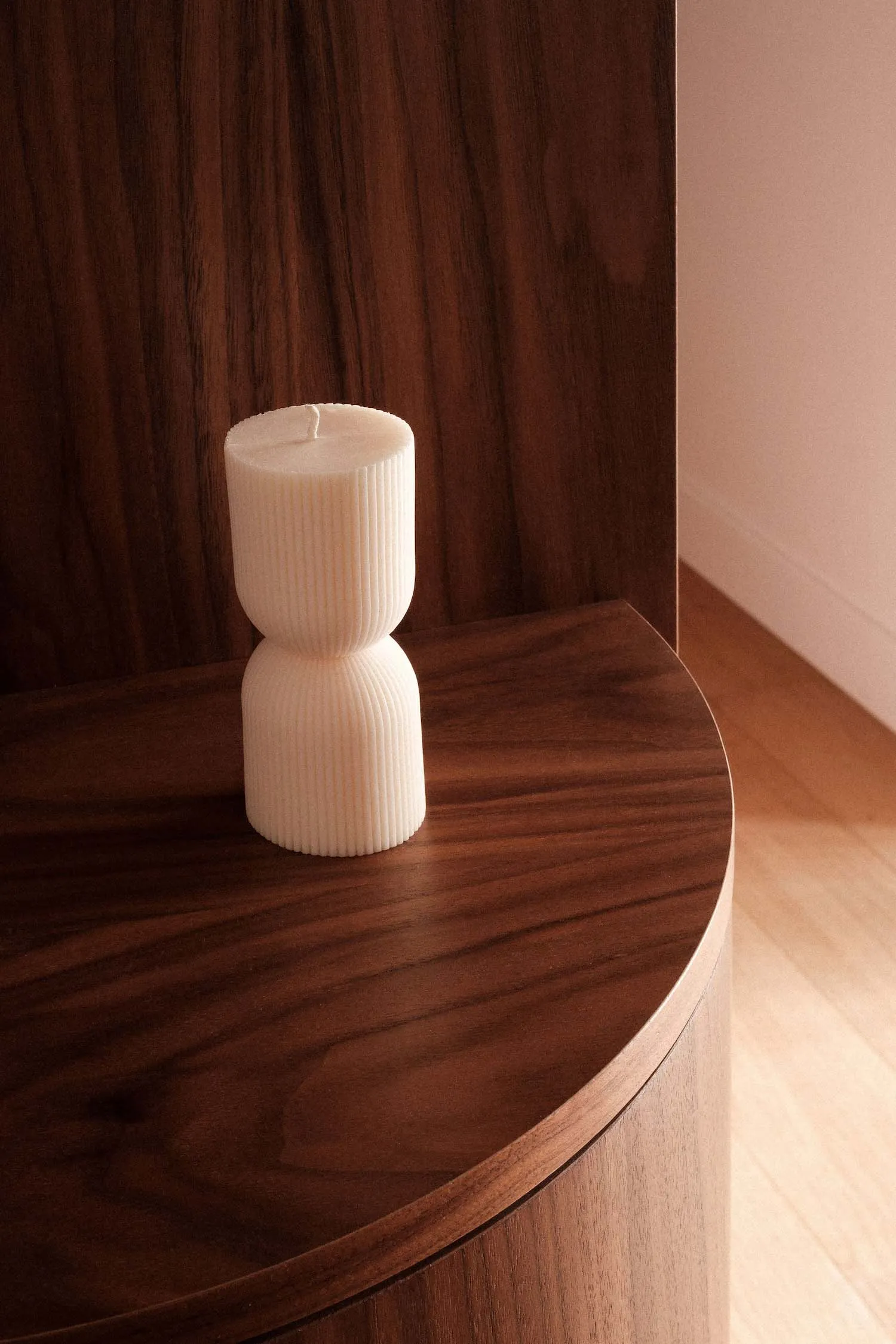 A white sculptural candle with ribbed texture sits on a wooden shelf.