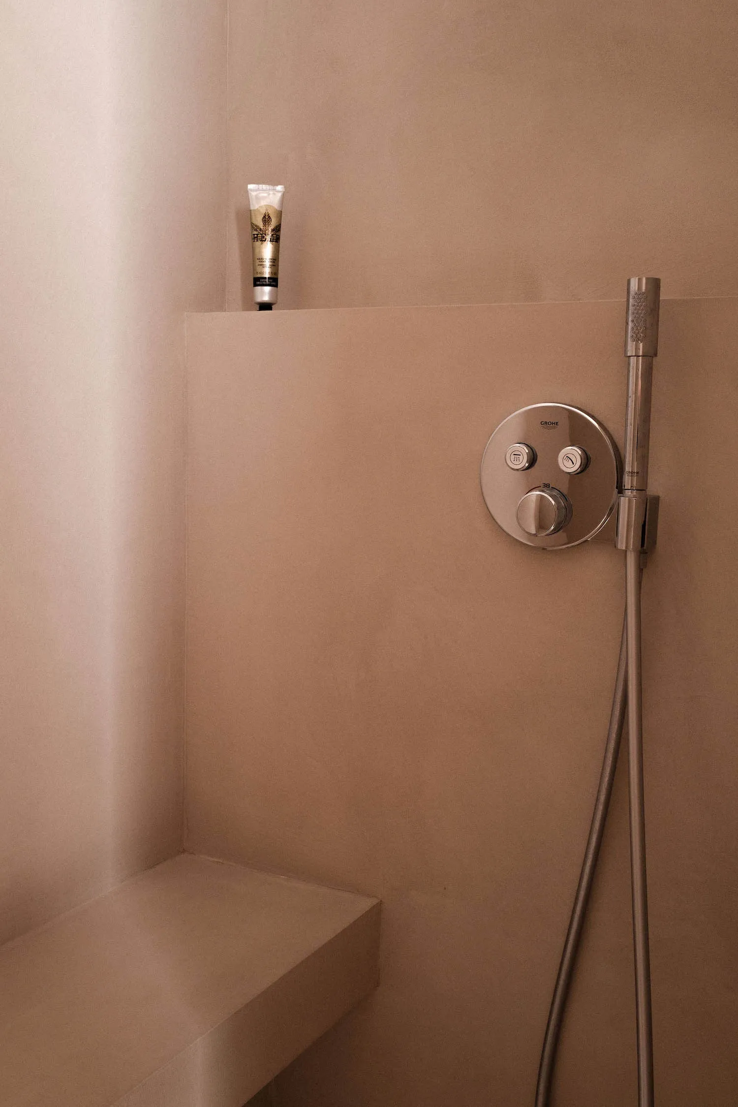 A minimalist bathroom with concrete walls, a hand shower, and a small bench.