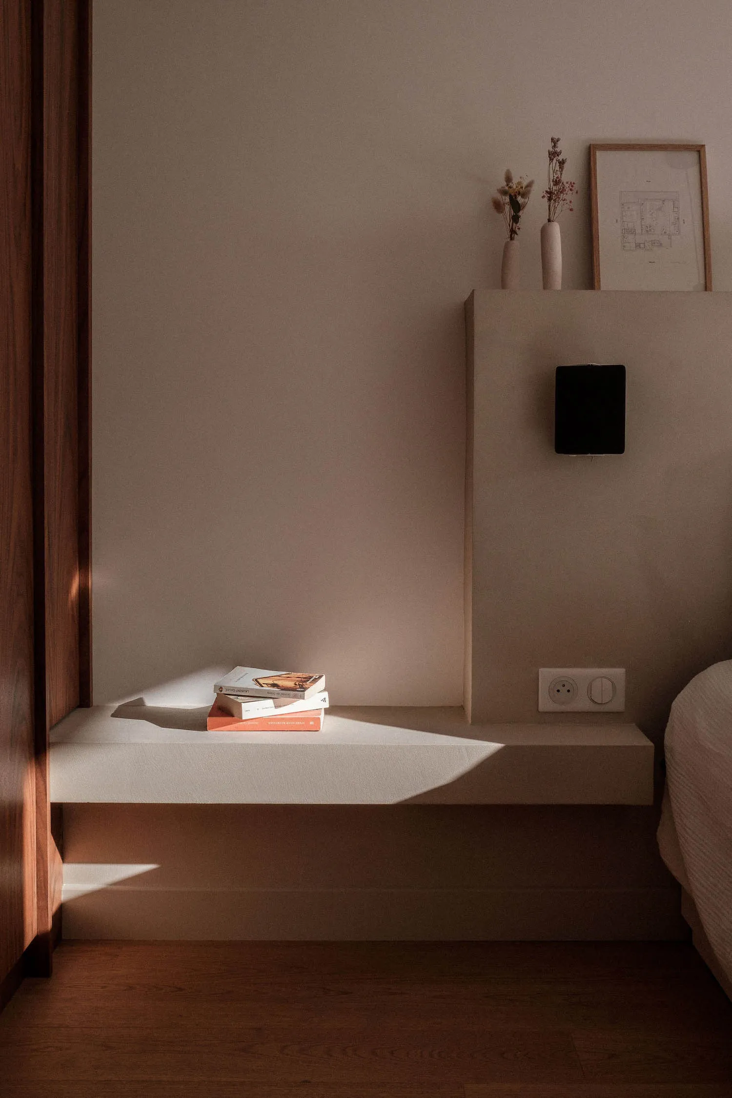 A minimalist bedroom with a modern design, featuring books on a floating shelf, dried flowers, and a sleek, contemporary bed.