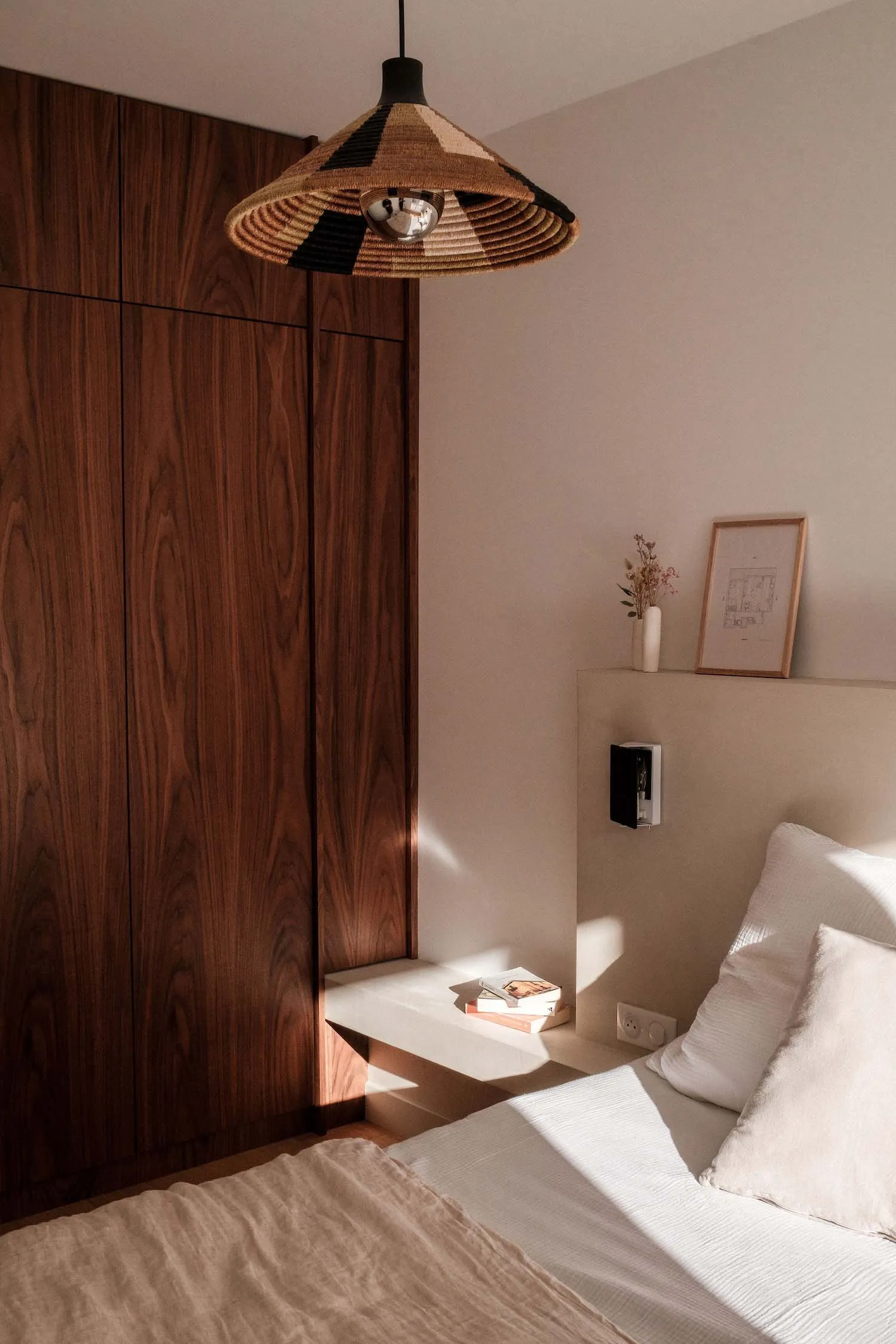 A minimalist bedroom featuring a wooden wardrobe, a bed with a plain white mattress and pillows, and a bedside table with a v