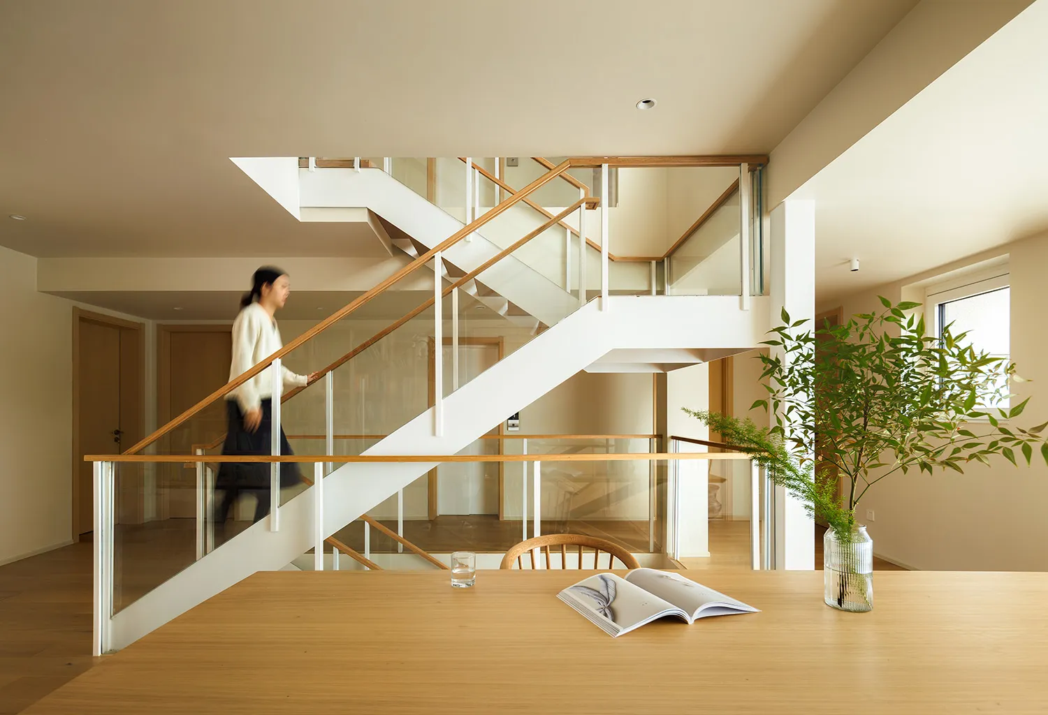 A modern living room featuring wooden and glass materials, with a person walking down a staircase in the background.