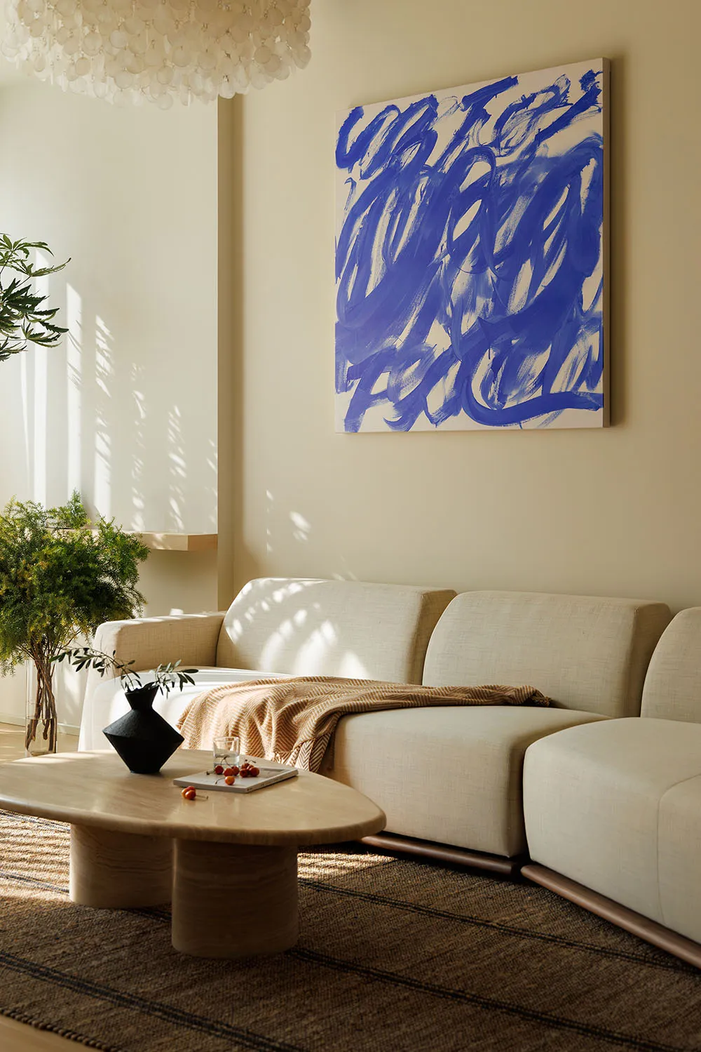 A modern living room with neutral-toned fabrics, a large abstract artwork, and a wooden coffee table with plants and a vase.