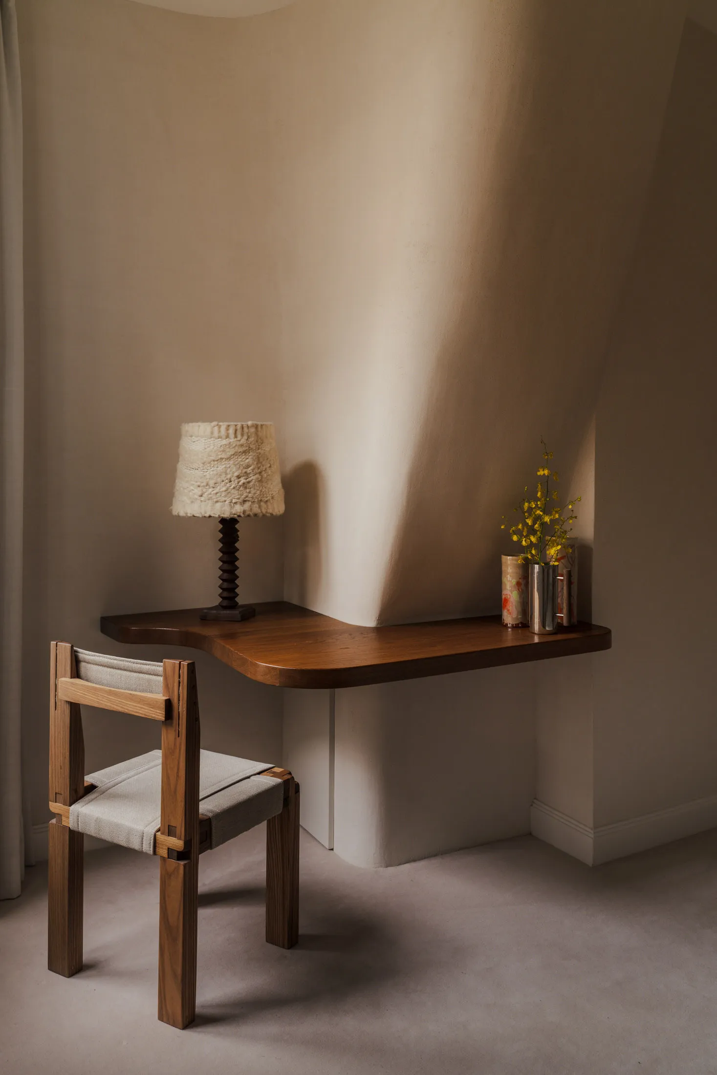 A modern home office setup featuring a wooden desk and chair, with a lamp and flowers on the desk.