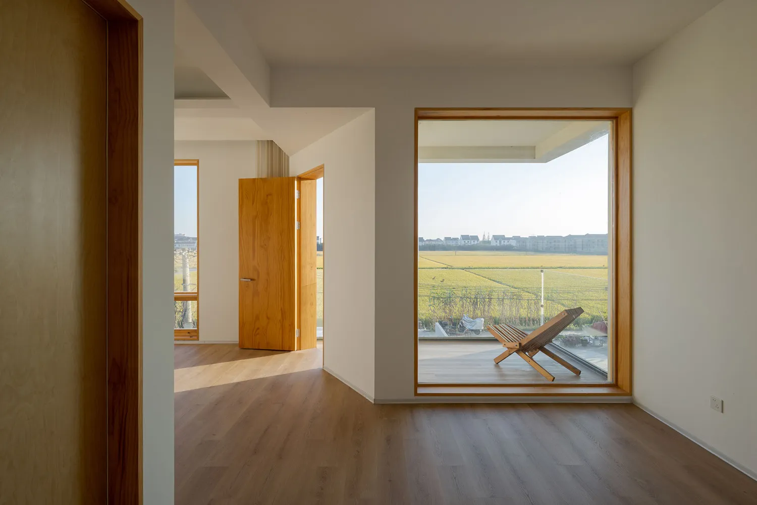 A modern residential space featuring a large window overlooking a field, with a wooden floor and a wooden chair.