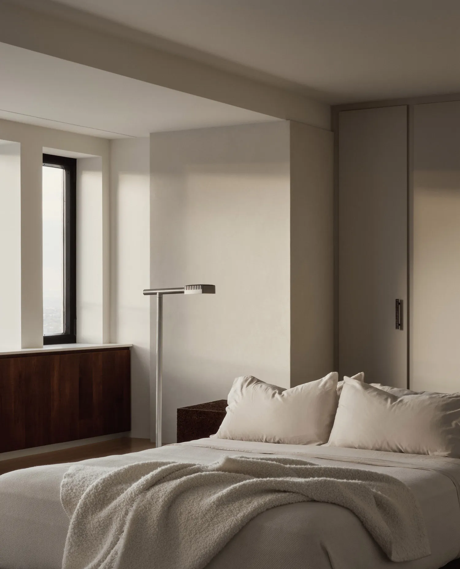 A minimalist bedroom with neutral-colored walls, bedding, and pillows, featuring a simple floor lamp and a window with a view