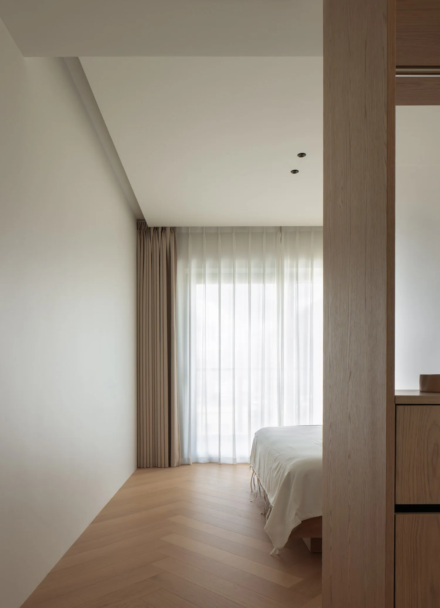 A modern minimalist bedroom with wooden flooring and light-colored curtains.