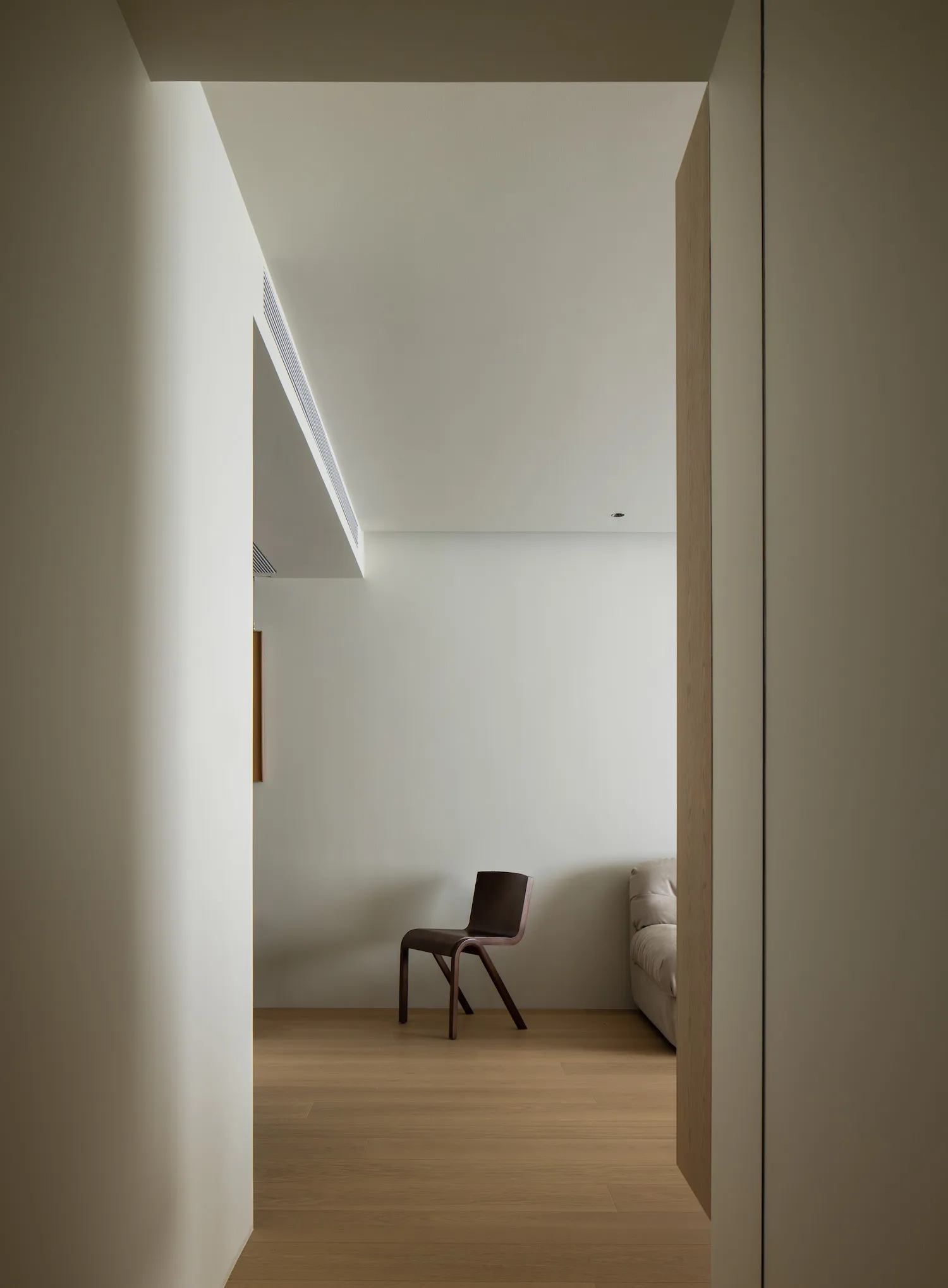 A minimalist living room with a wooden floor, a single dark wooden chair, and a light-colored sofa.