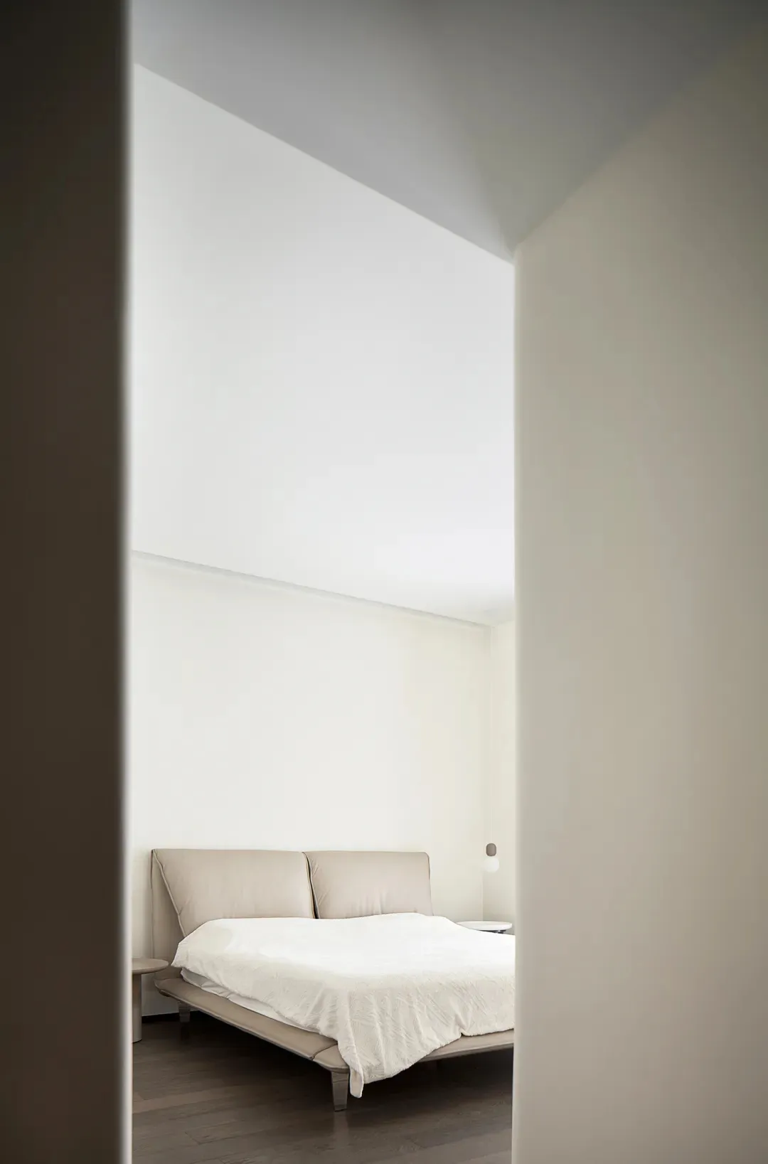 A serene bedroom with minimalist design, featuring neutral-toned furniture and a white bedspread, photographed from a doorway