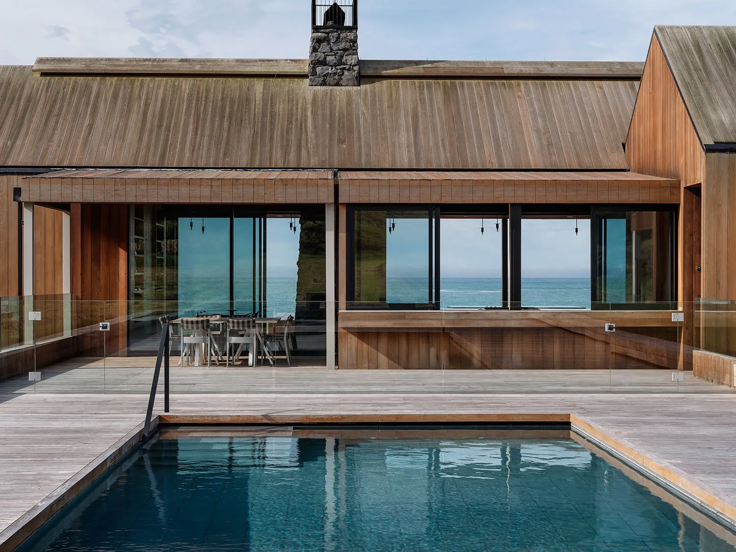 A modern minimalist house with a wooden exterior, large windows overlooking the ocean, and a swimming pool.