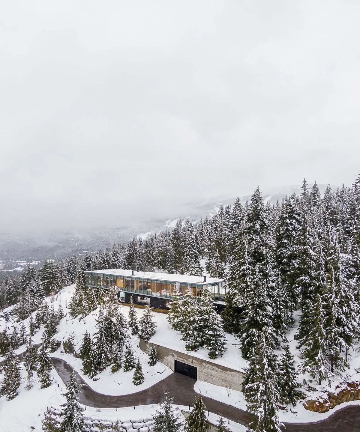 A modern, glass-fronted residence built on a snowy, forested hillside.