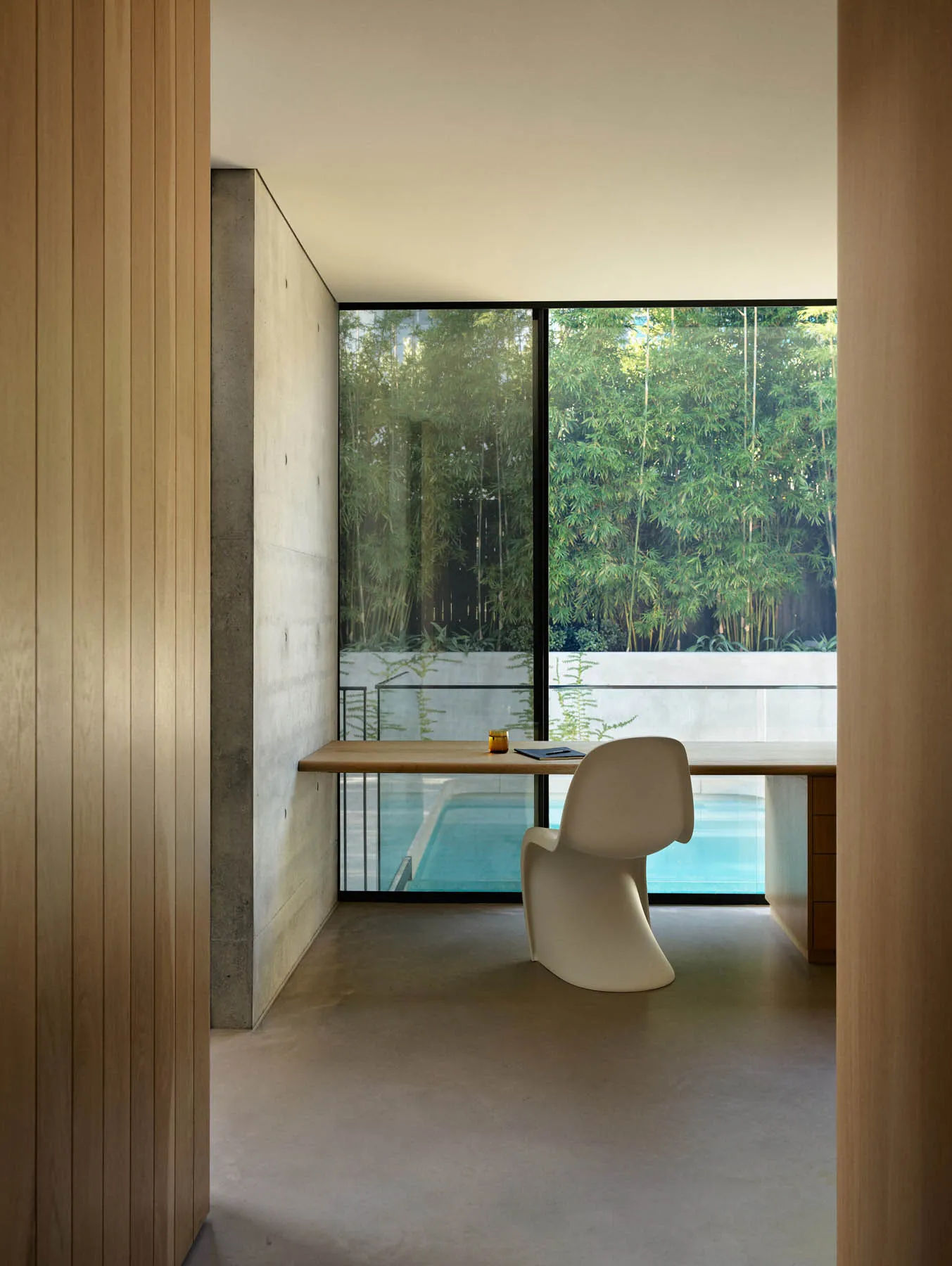 Minimalist office space featuring a concrete desk and chair with a large window overlooking a pool and greenery.