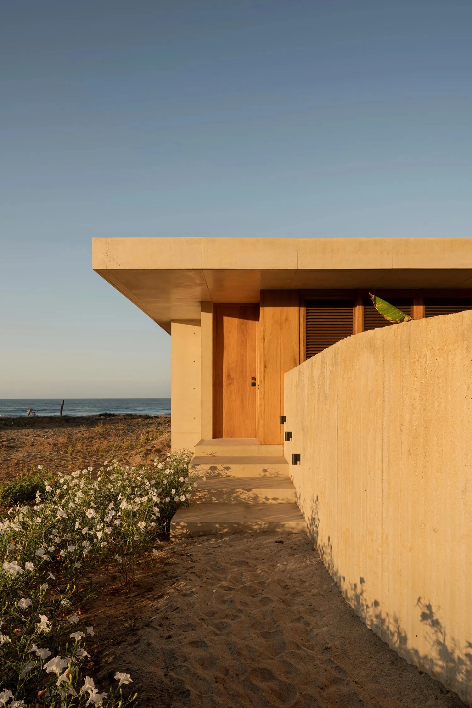 Modern minimalist residential exterior with concrete walls and a wooden door on a sandy beach.