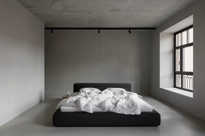 A minimalist bedroom featuring a grey concrete bed frame with white bedding, set against a grey concrete wall and floor.