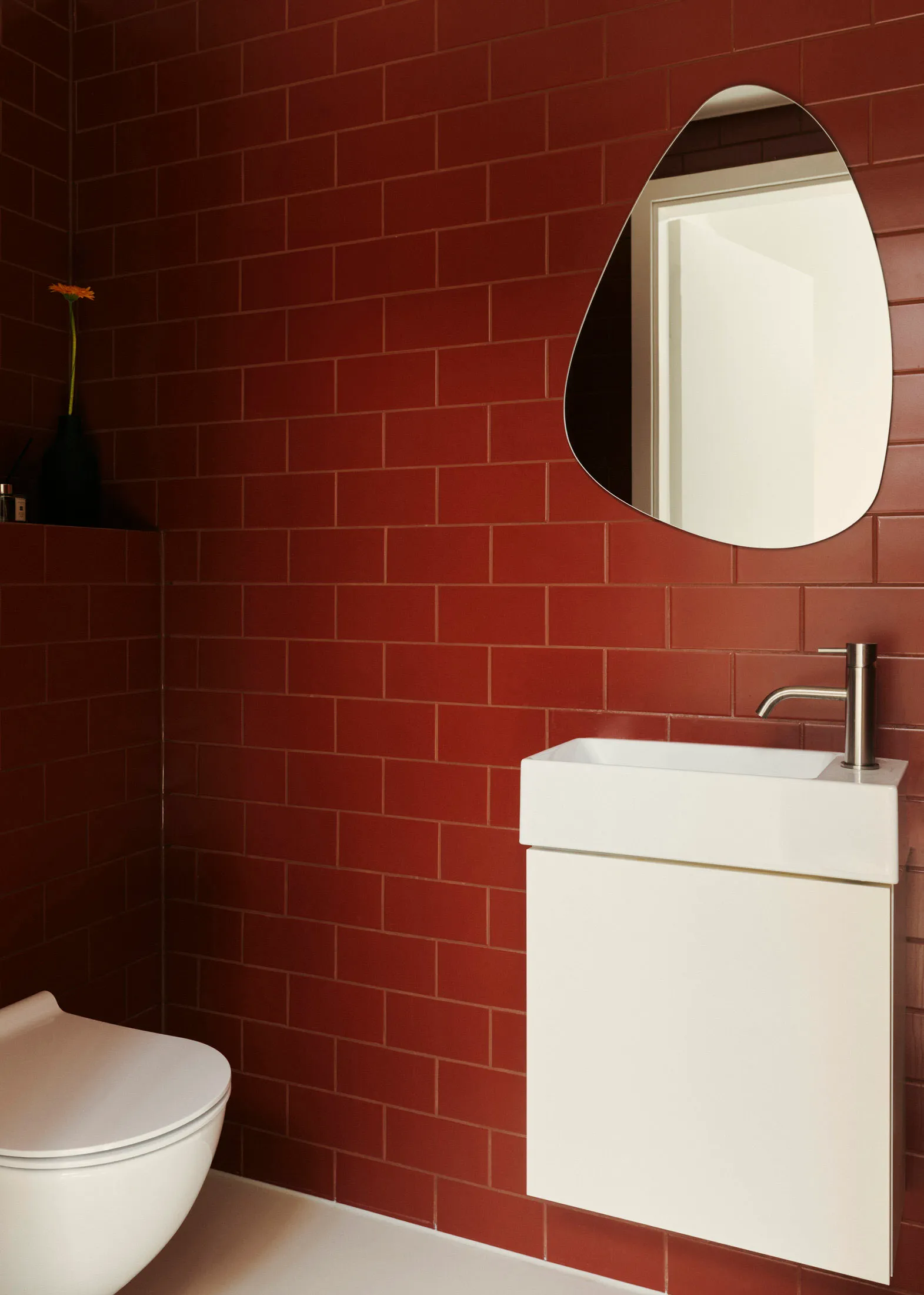 Modern minimalist bathroom with red ceramic tiles, white fixtures, and a single vase with flowers on the shelf.