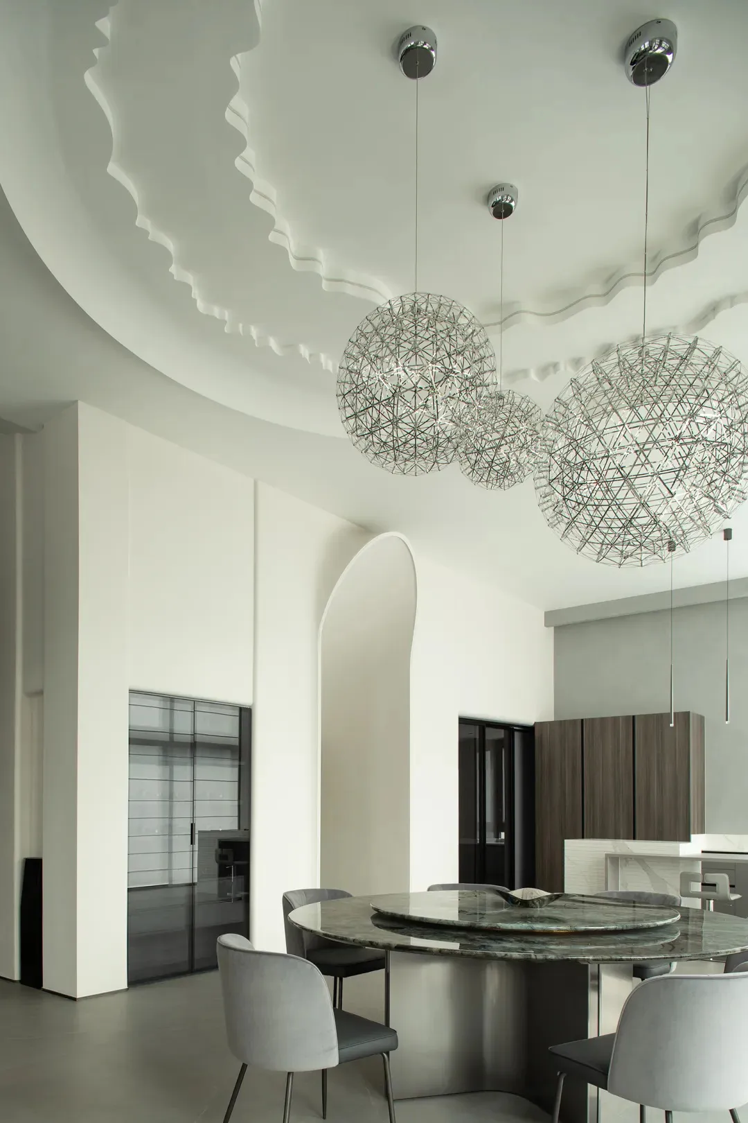Modern dining room featuring a metal chandelier with geometric patterns, grey upholstered chairs, and a round granite table.