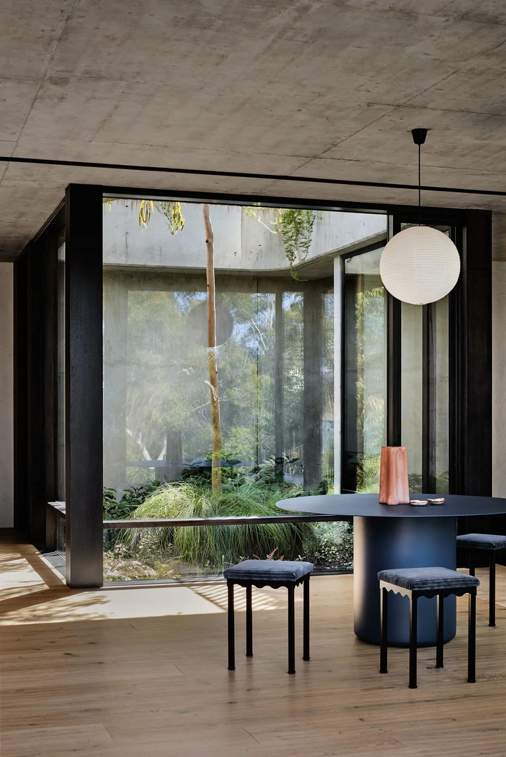 A modern, minimalist dining room with a large round table, stools, and a concrete and glass structure visible through large w