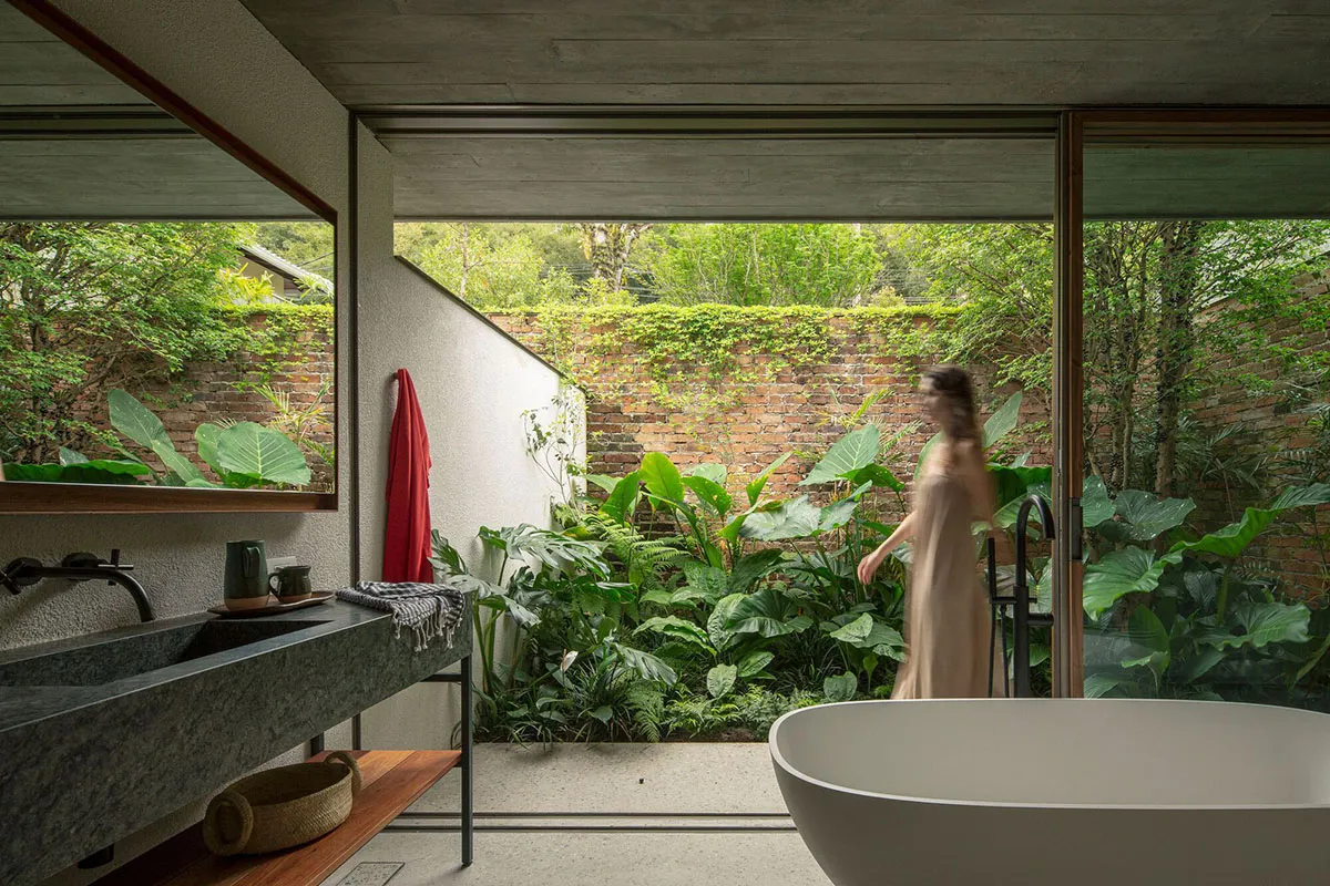 Modern bathroom with concrete walls, large windows, and green tropical plants outside, featuring a freestanding white bathtub