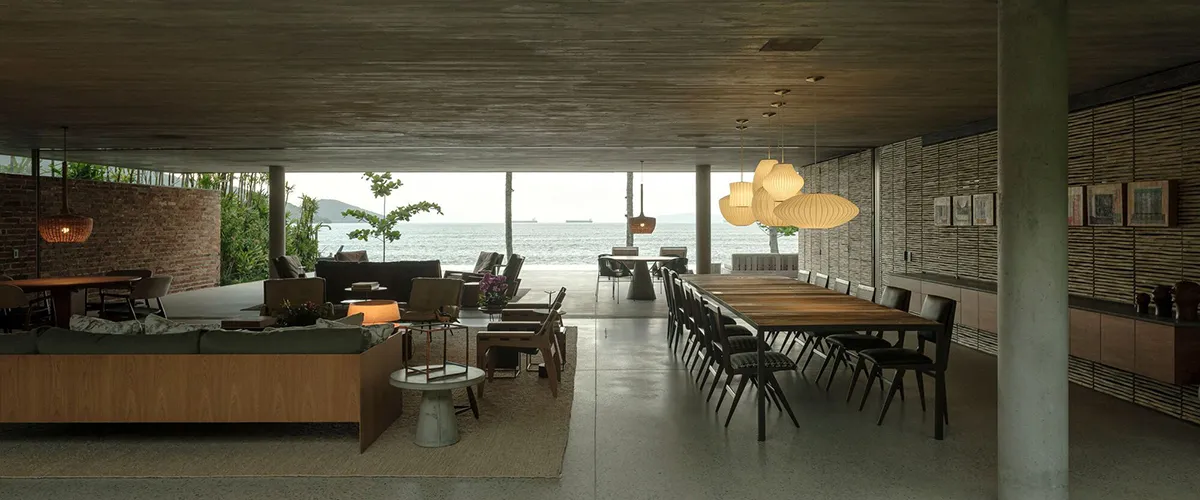 Modern, open-plan living room with concrete walls and floor, large windows overlooking the sea, and ergonomic furniture.