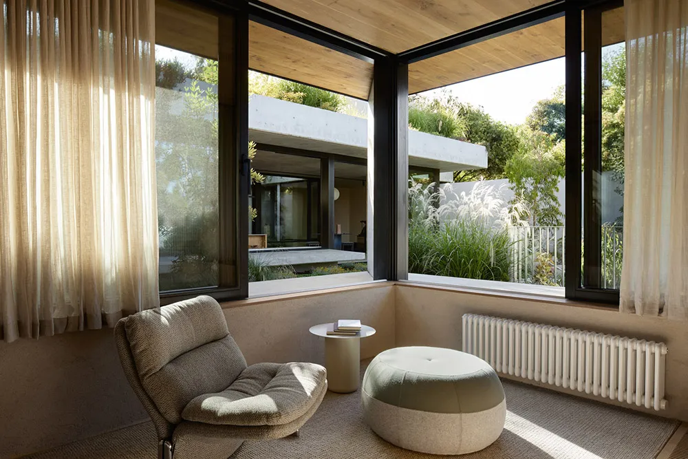 A modern minimalist living space with a comfortable armchair, round ottoman, and large windows allowing natural light in, ove