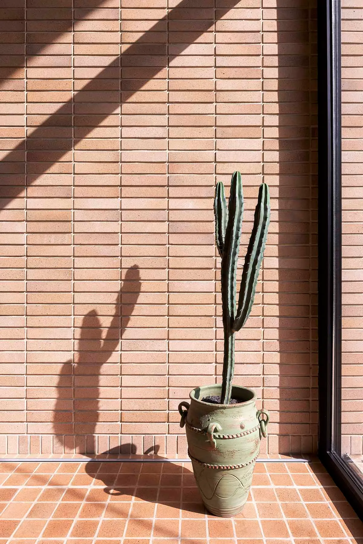 A cactus plant in a green ceramic pot on a tiled floor against a brick wall.