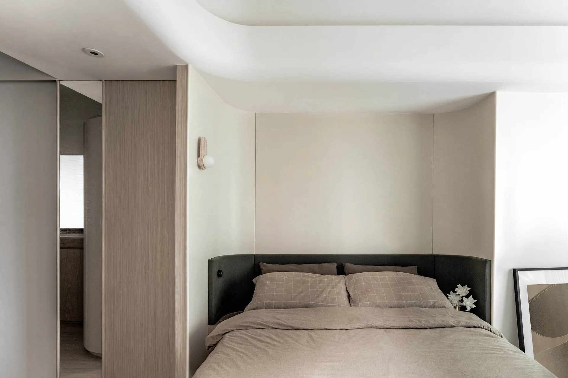 A minimalist bedroom featuring a bed with a black upholstered headboard and beige bedding, alongside a wooden headboard panel