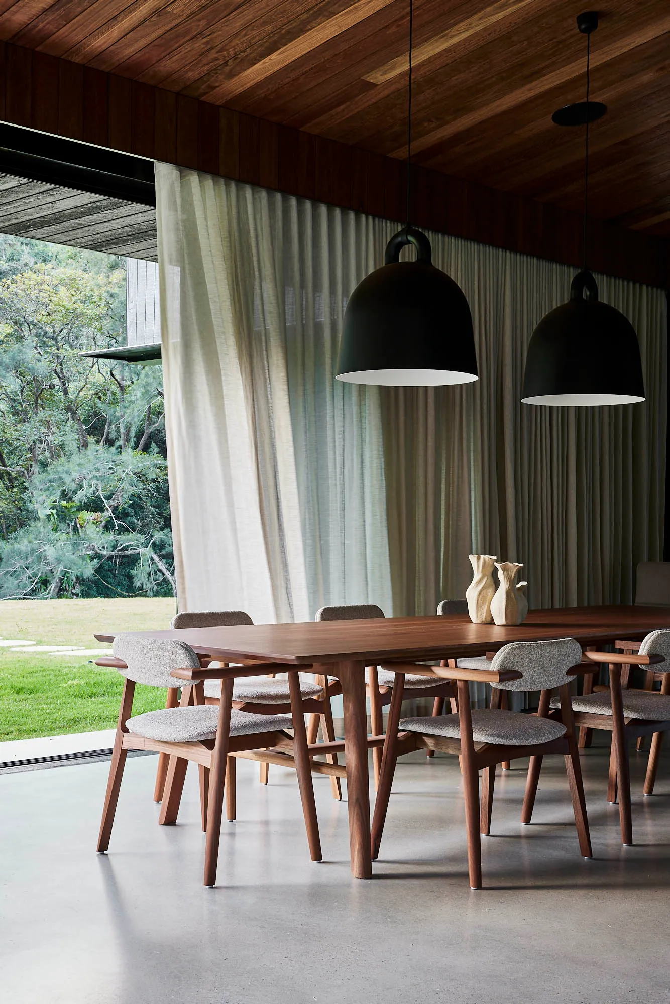 A modern dining room with wooden furniture and concrete flooring, featuring a large table and chairs, large windows with curt