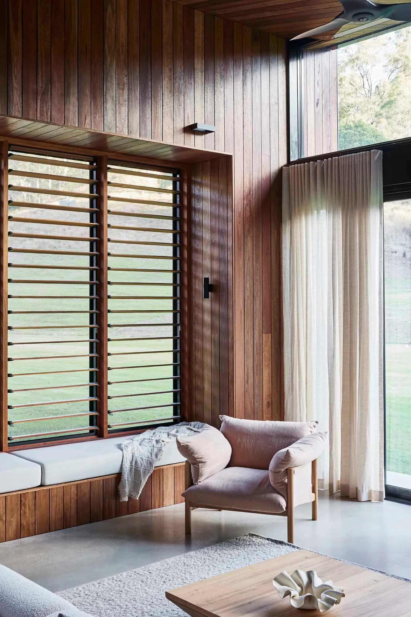 A modern living room featuring wooden furniture and large windows with wood-patterned blinds.