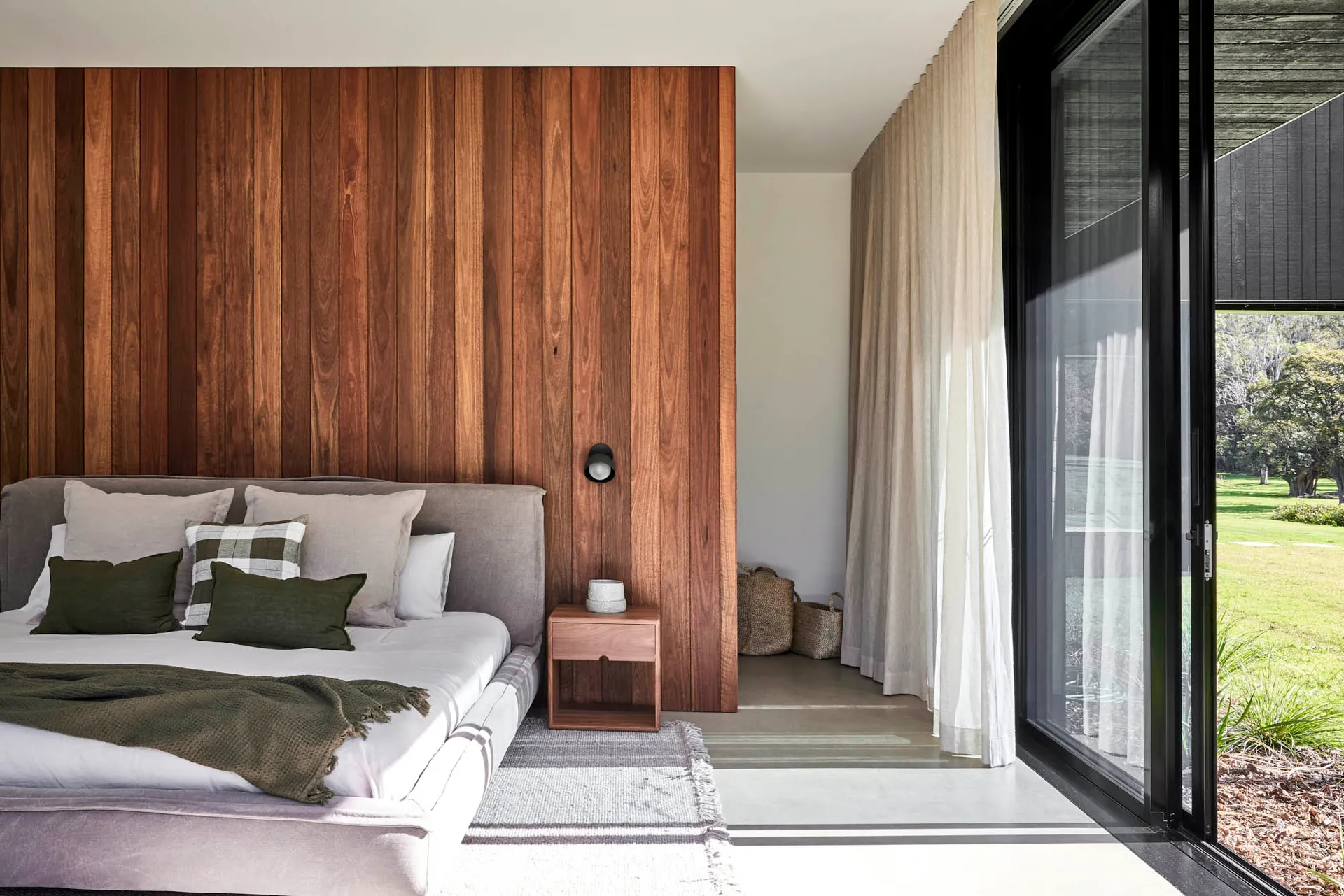 A modern minimalist bedroom with wooden panels on one wall, concrete flooring, and a view through a glass door to an outdoor