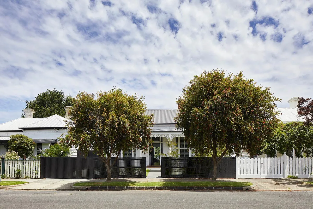 A white Victorian-style house with a brick fence and trees in front of it, set against a blue sky with white clouds.