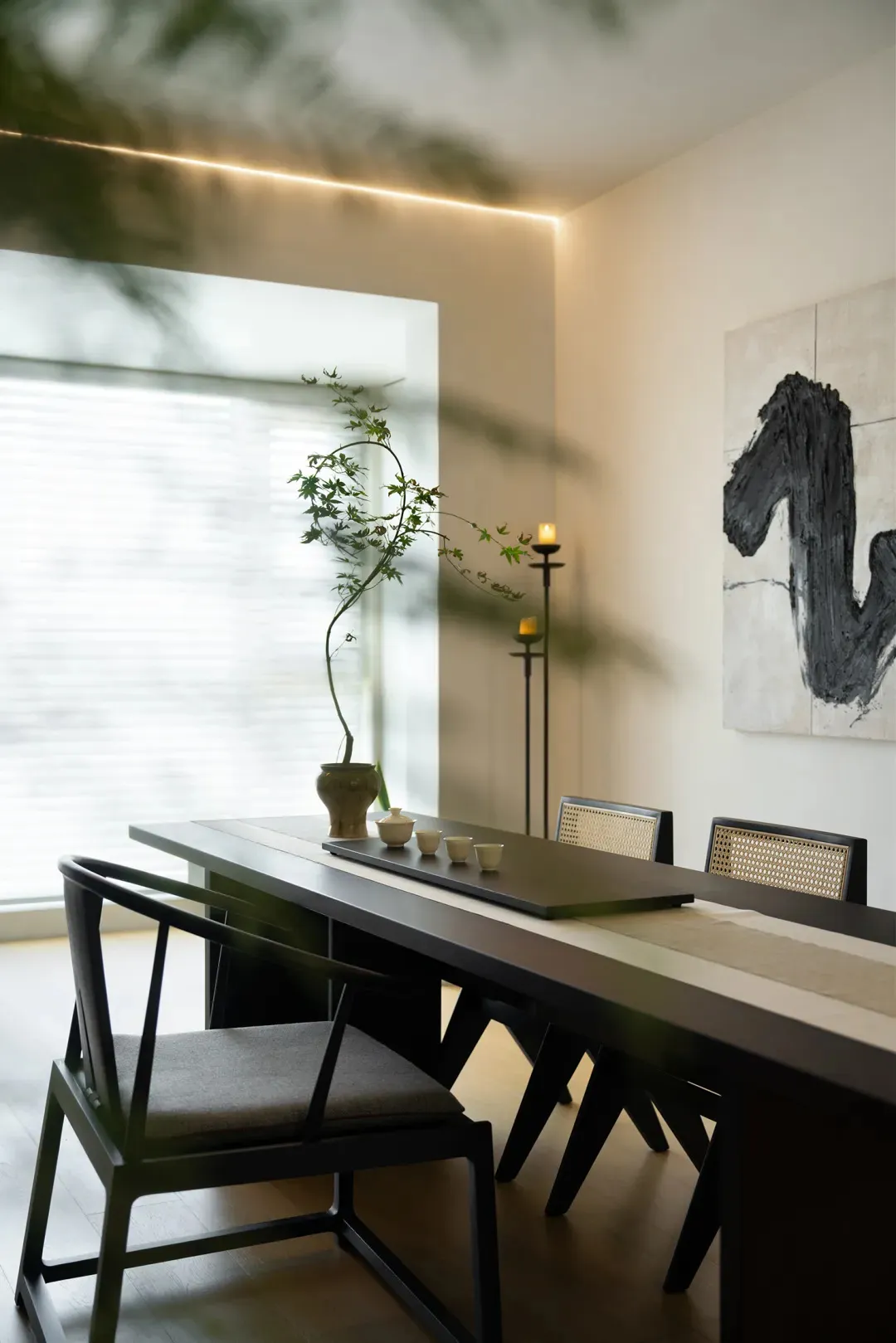 A minimalist dining room with a long wooden table and chairs, placed near a large window with a plant and candles.