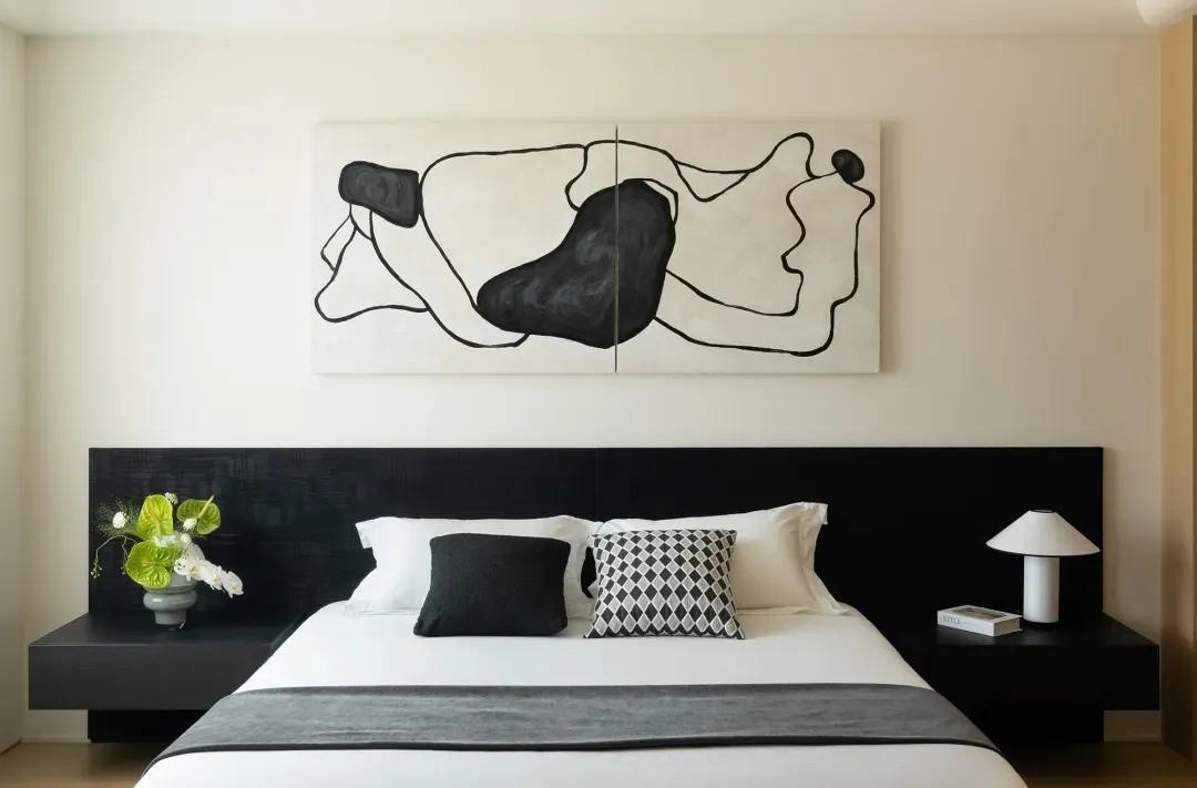 A minimalist bedroom with a textured wooden headboard and white bedding. A black and white abstract painting hangs above the