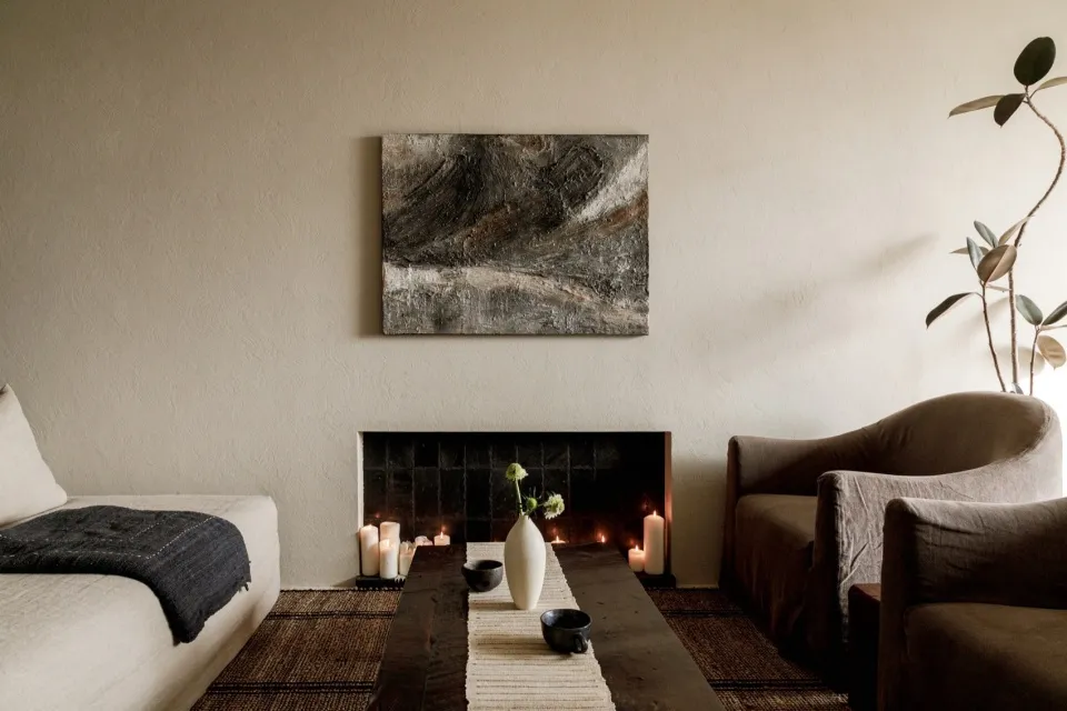 A minimalist living room featuring a wooden fireplace with lit candles, a vase with flowers, and a dark wooden coffee table.
