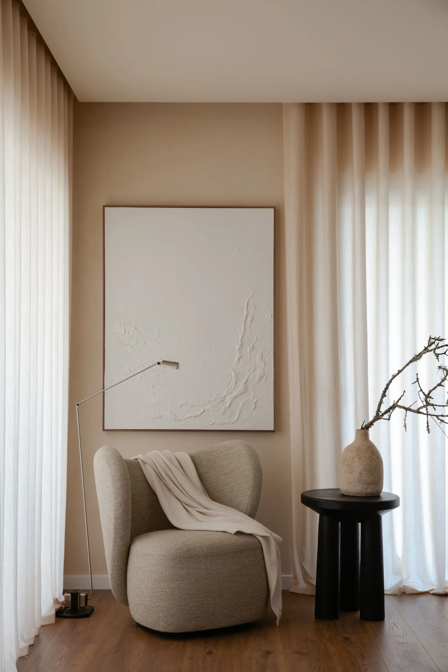A minimalist living room featuring a beige armchair with a blanket, a black side table with a vase, and a textured wall art p