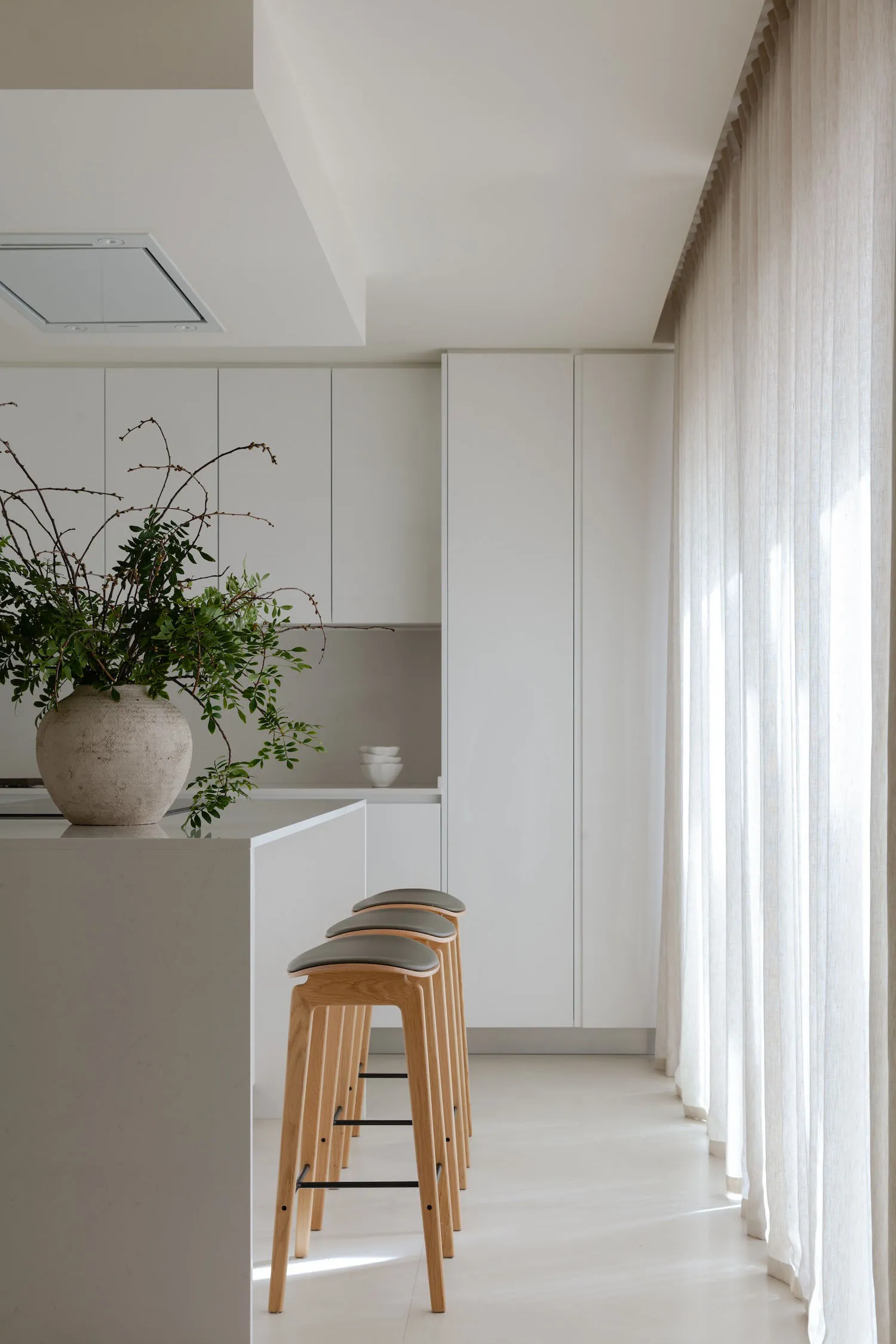 A minimalistic white kitchen with clean lines, modern stools, and large windows.