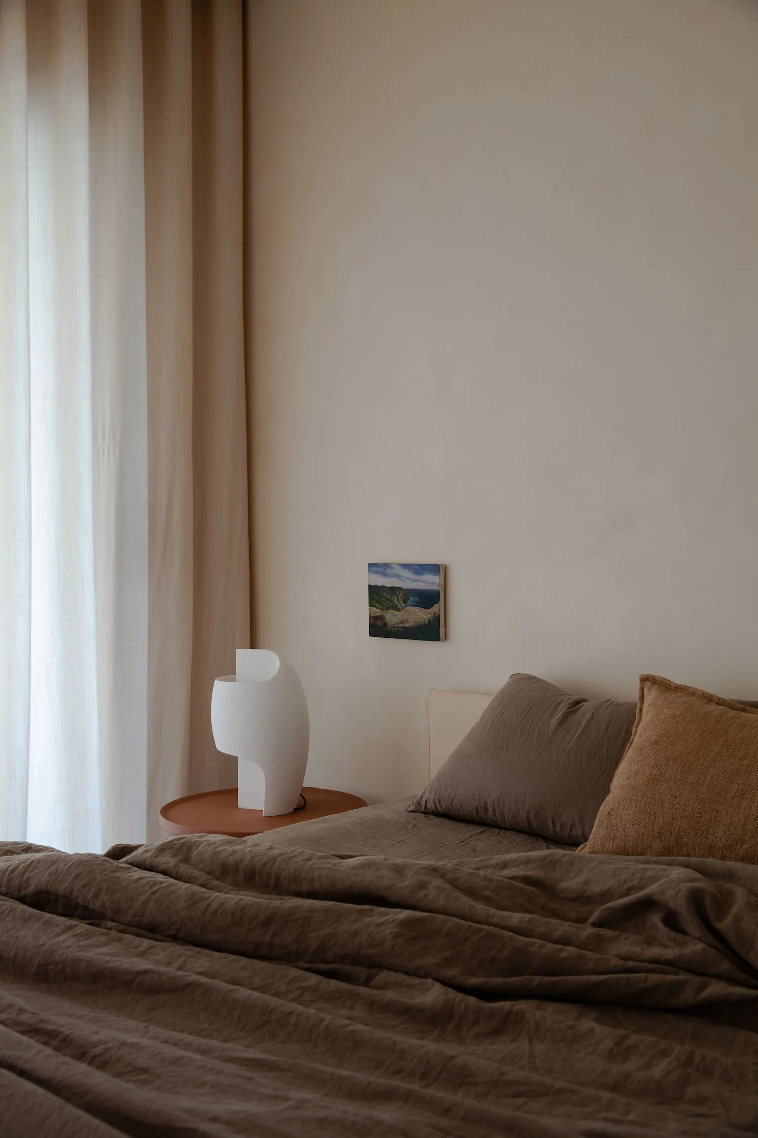 A modern bedroom featuring a bed with brown textiles and a side table with a sculptural lamp.