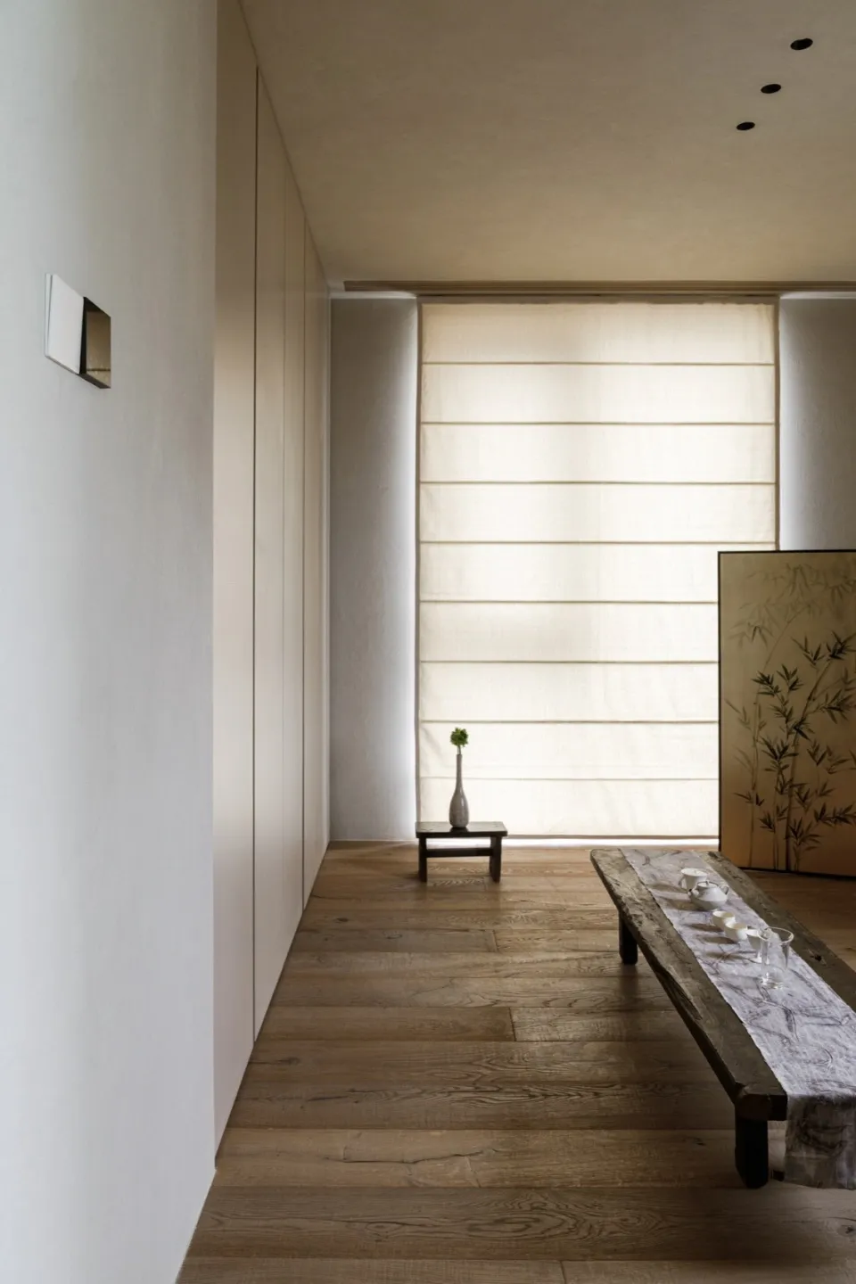 A modern minimalist living room with wooden furniture, a small table with a vase, and a large window with blinds.