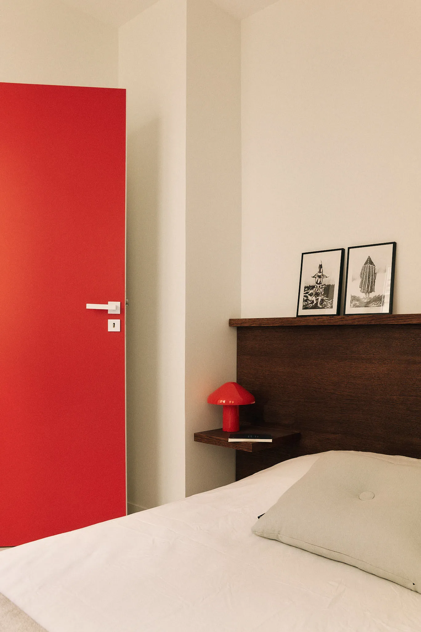 Photograph of a minimalist bedroom featuring a red door, wooden headboard, and a white pillow.